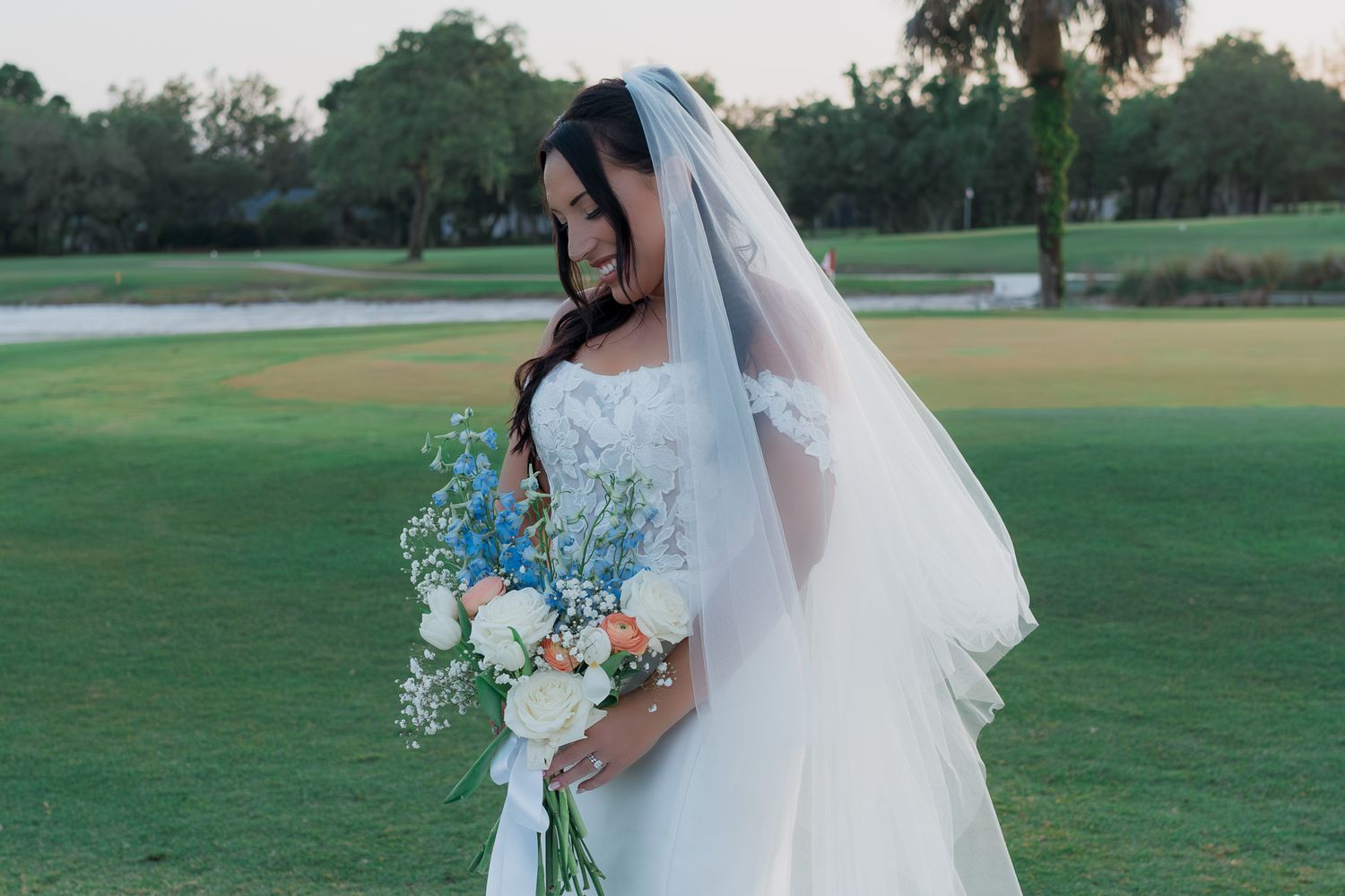 Bride in white gown and veil holding blue and white floral bouquet on lush green golf course.