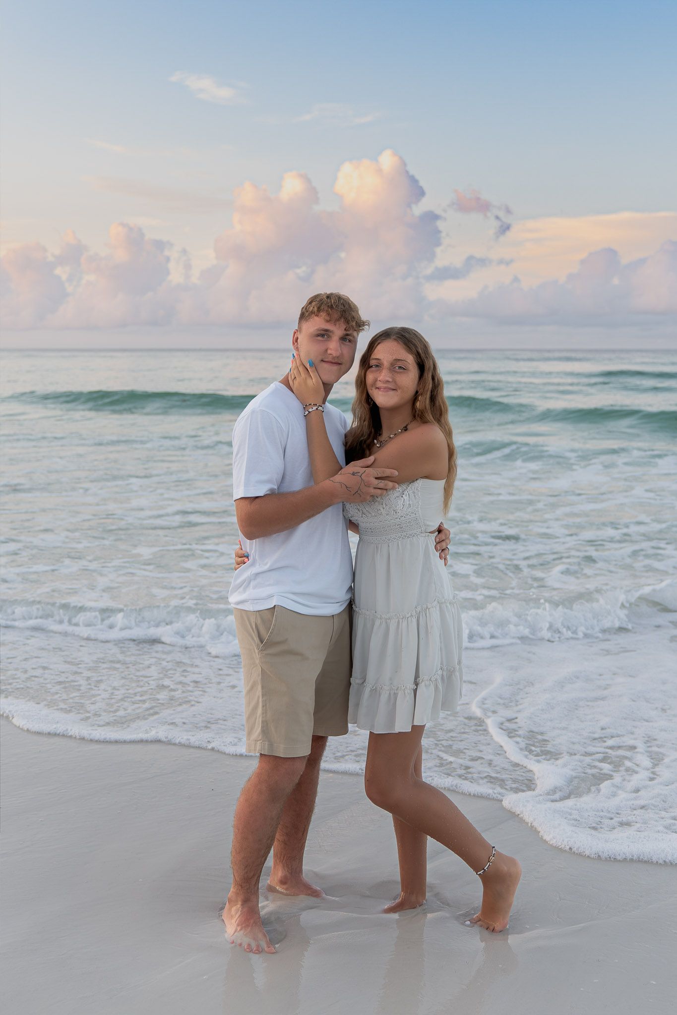Young couple embracing on a sandy beach at sunset with turquoise waves and pastel clouds behind them.