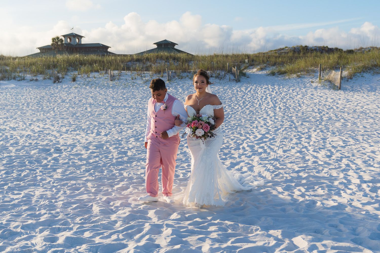 Couple in wedding attire on white sand beach, bride in mermaid gown holding bouquet, groom in pink suit.