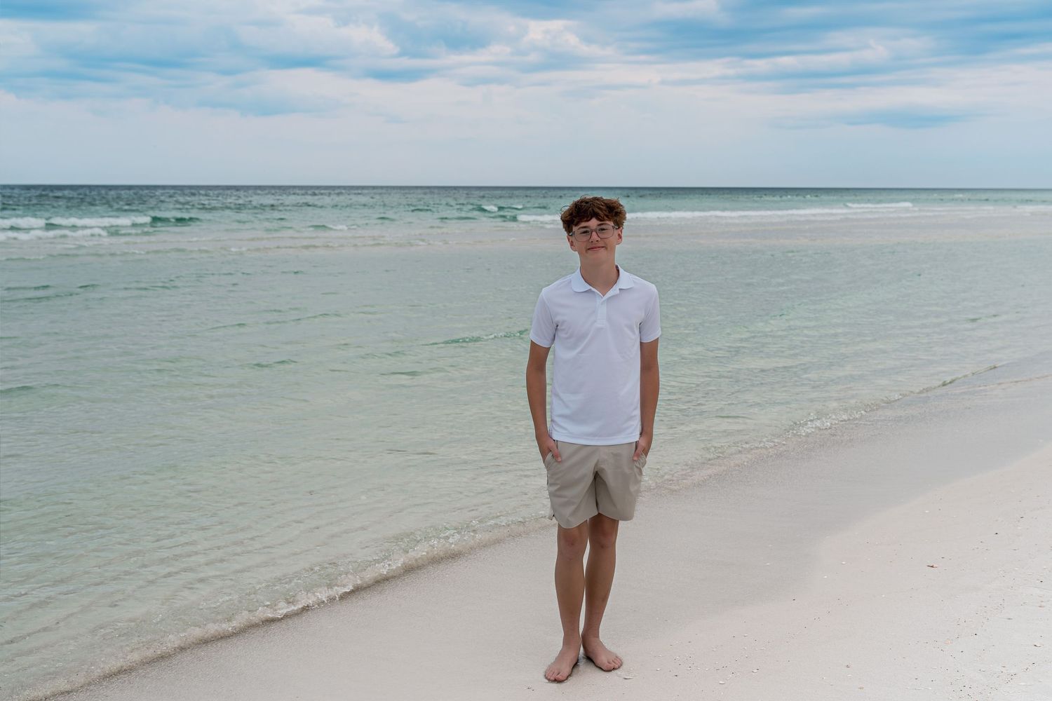 Young man in white polo and khaki shorts standing barefoot on a white sandy beach with turquoise ocean water.