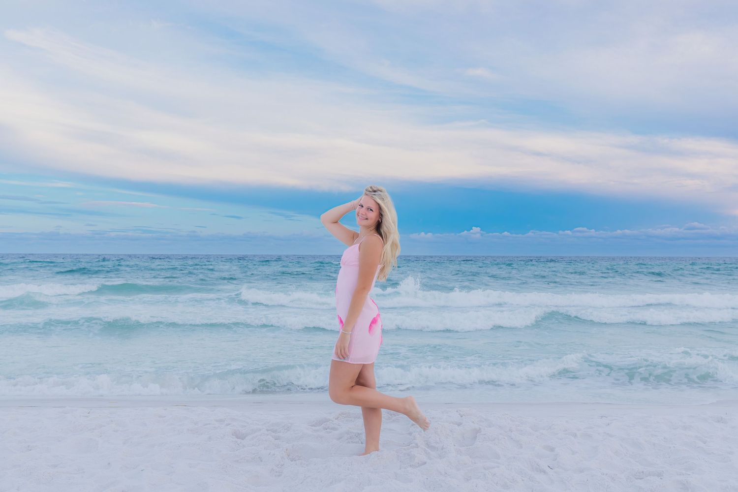 Blonde woman in pink dress posing on a sandy beach with turquoise waves and pastel sky.