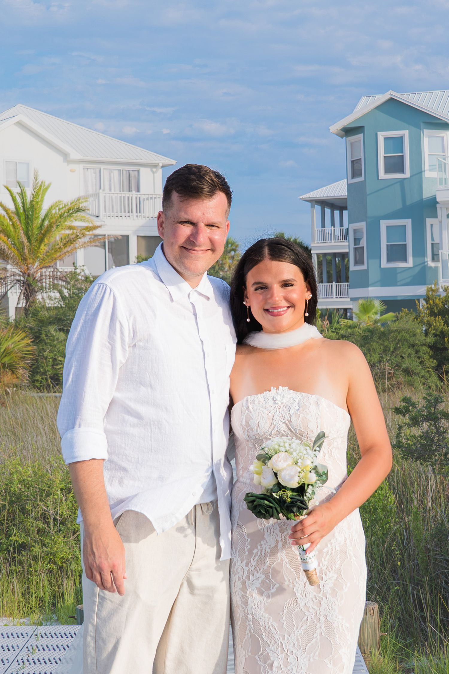 Smiling couple in white wedding attire poses outdoors near beach houses, bride holding white flower bouquet on sunny day.