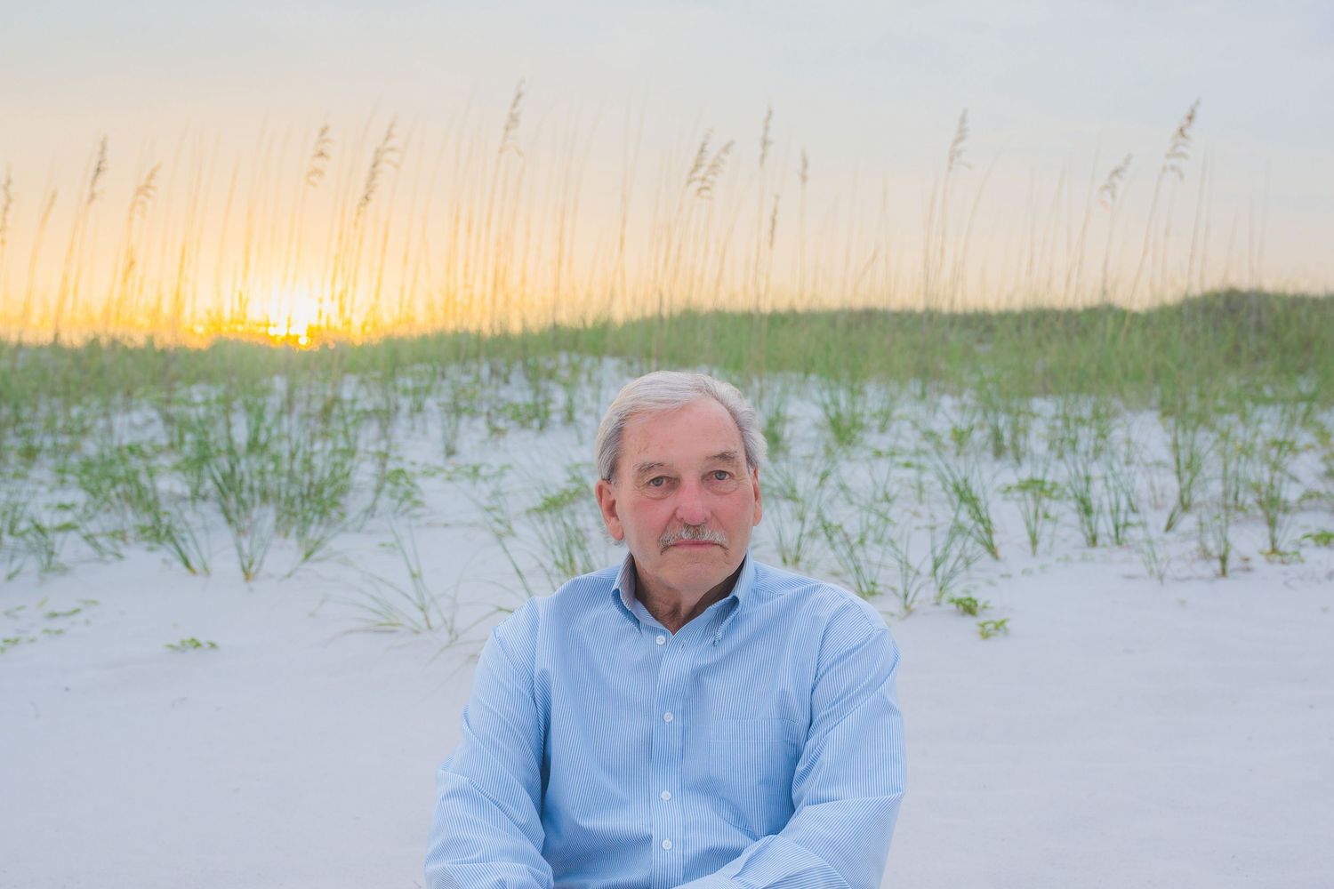 Man sitting on white sand beach at sunset with sea oats and golden sky behind him.