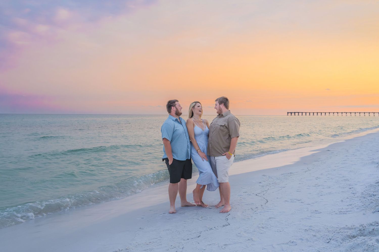 Three people pose together on a white sand beach at sunset with a colorful pink and orange sky and pier in background.