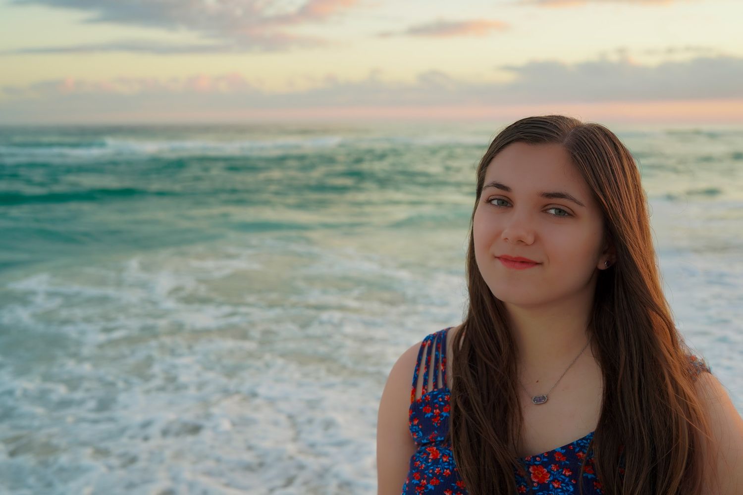 Young woman with long brown hair smiles at the beach during a colorful ocean sunset portrait session.