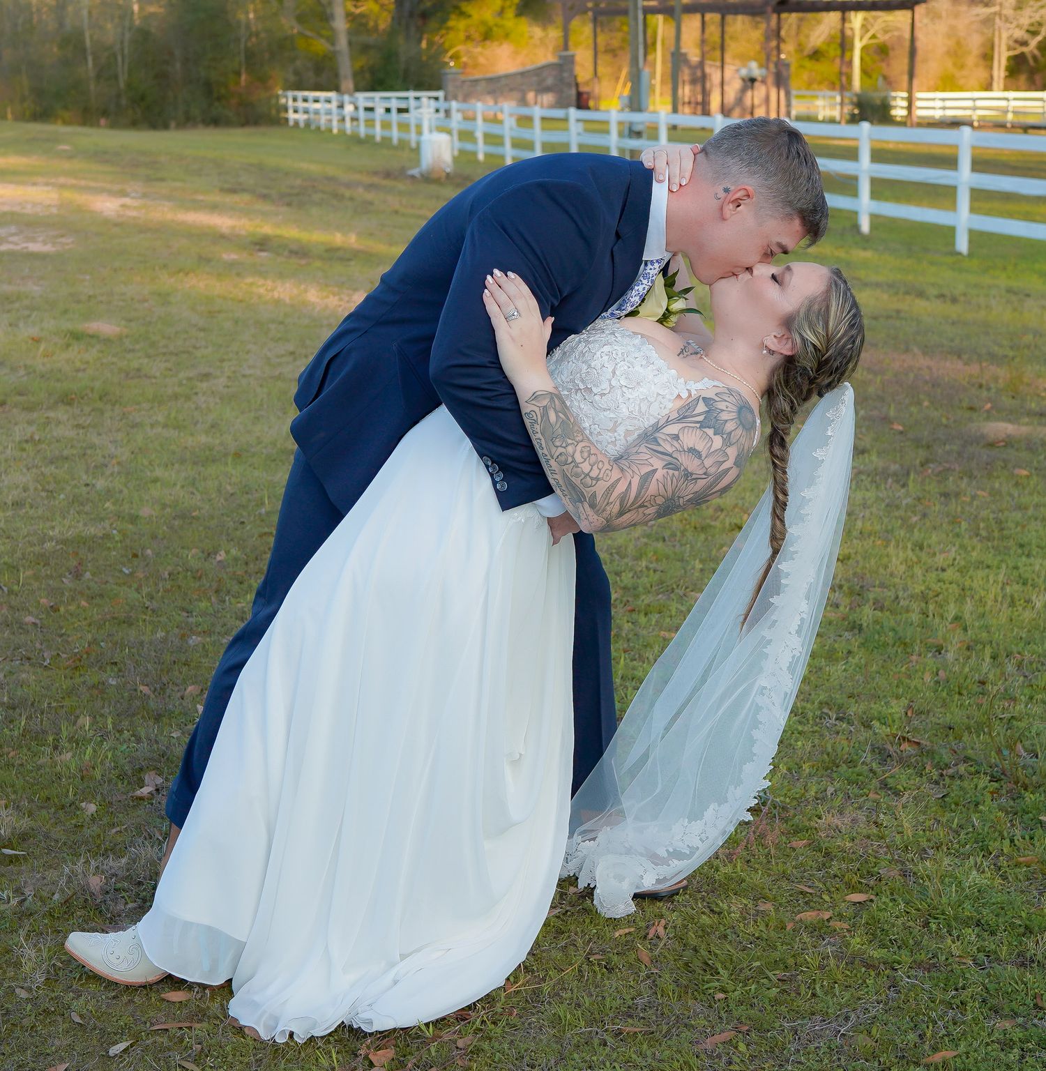 Groom dips bride for a romantic kiss outdoors near white fence during wedding portrait session.