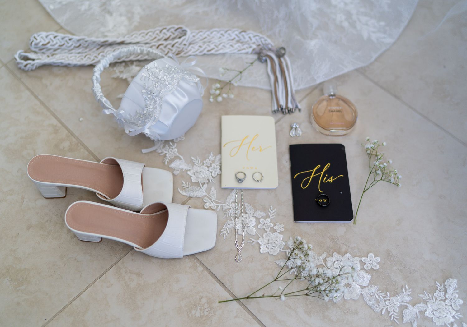 Elegant bridal flat lay with white heels, lace veil, purse, perfume, earrings, and His vow books on tile floor.