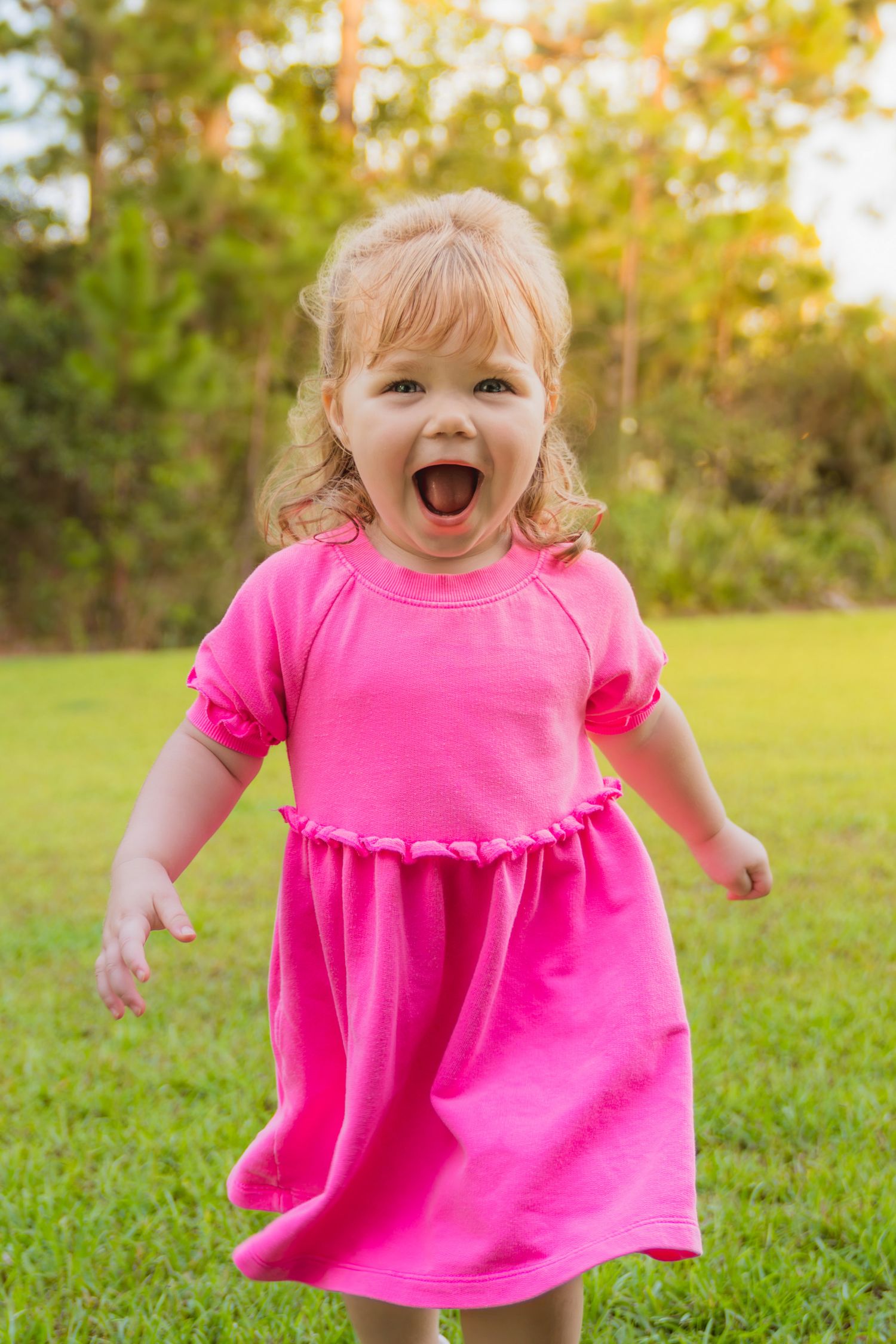 An excited toddler girl in a bright pink dress laughs with arms outstretched on a green lawn with trees behind her.
