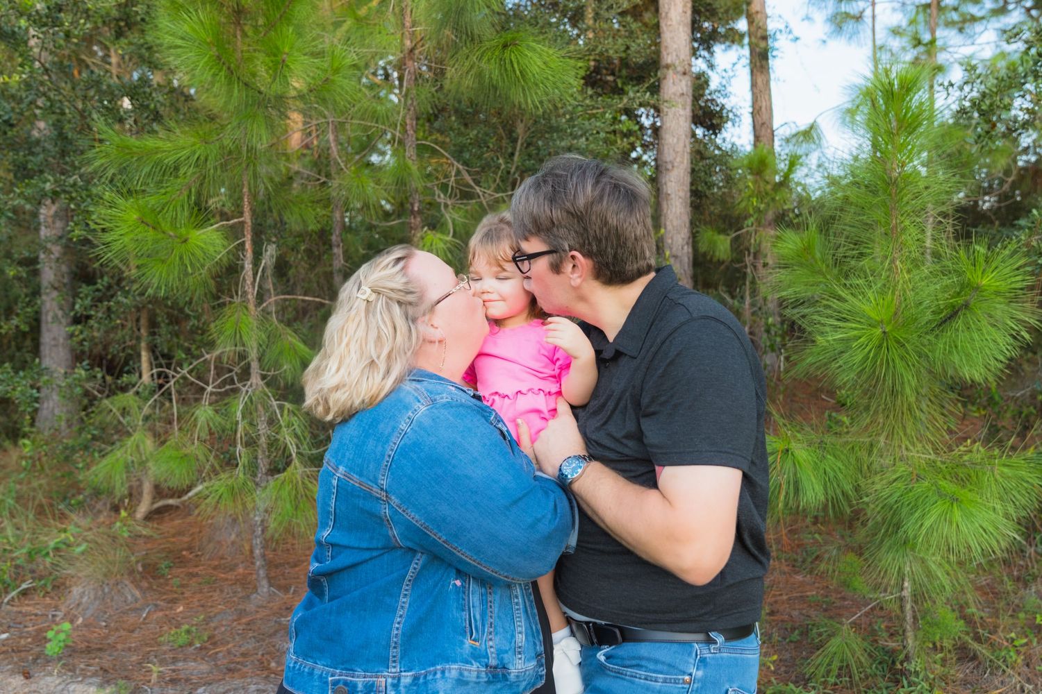 Two parents kissing their newborn baby outdoors among tall green pine trees in a heartwarming family portrait.