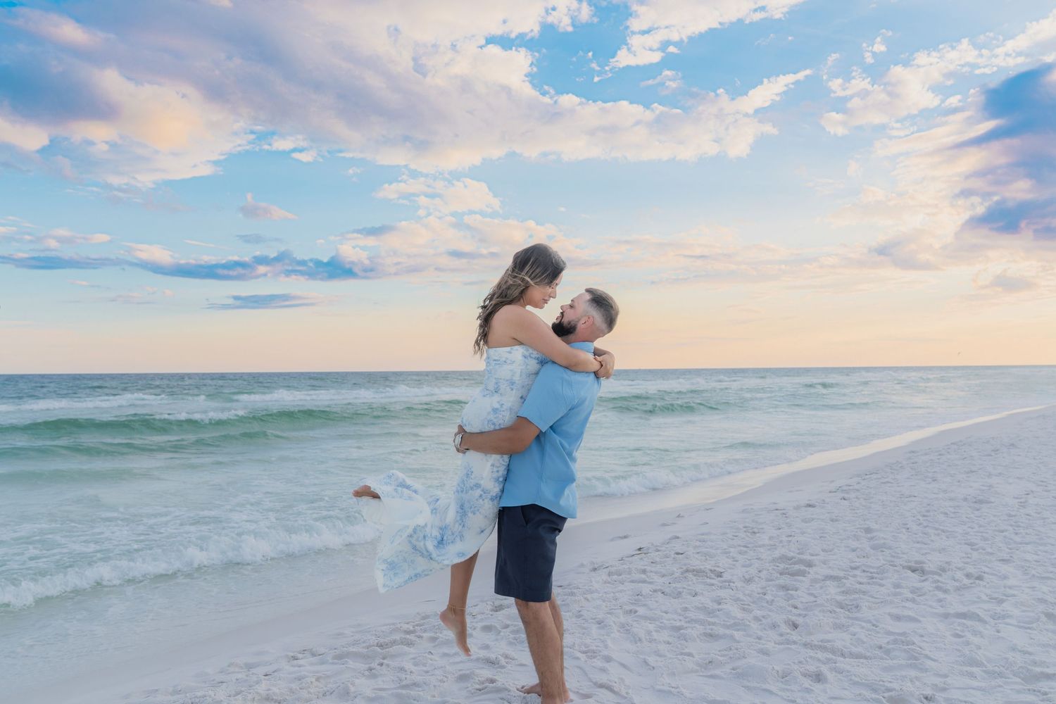 Couple embracing on a white sand beach at sunset with turquoise ocean waves and pastel sky backdrop.