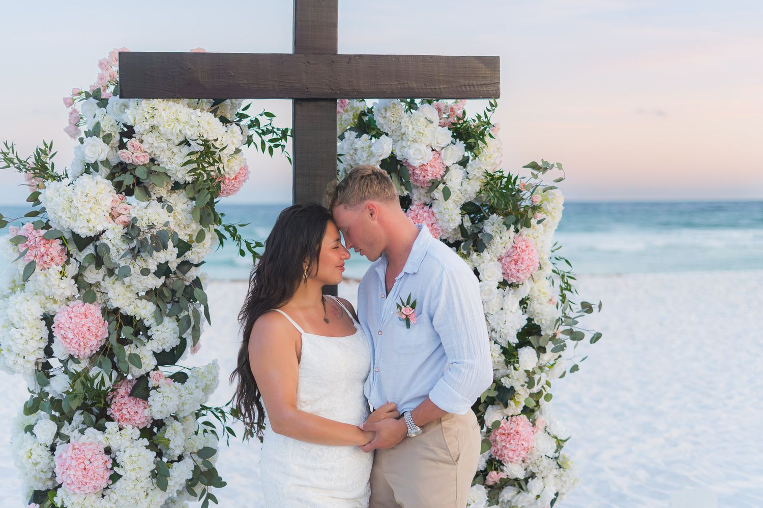 Couple sharing romantic moment under floral cross arch on beach at sunset, surrounded by pink and white flowers.