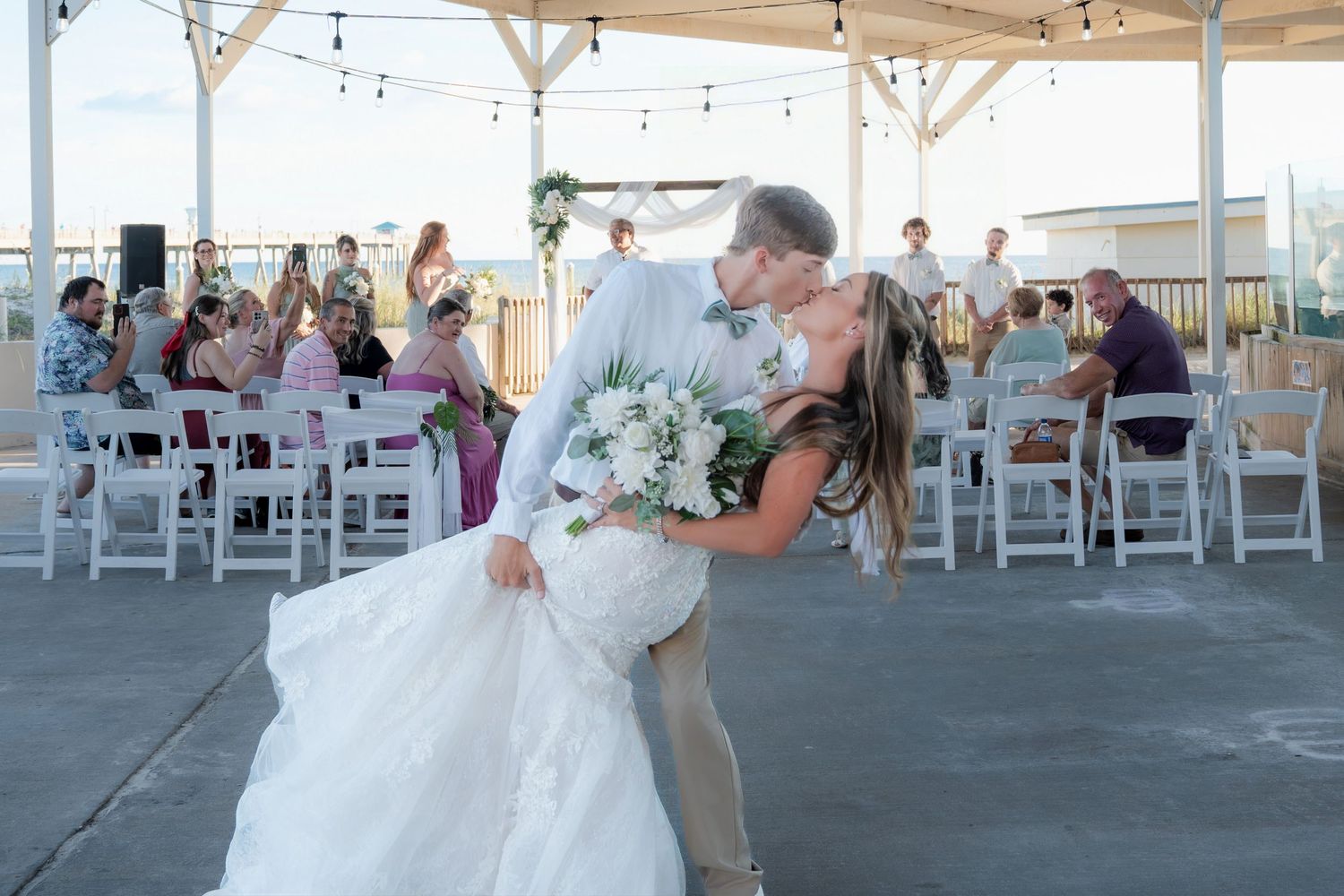 Bride and groom share romantic dip kiss on outdoor wedding reception dance floor with guests seated around them.