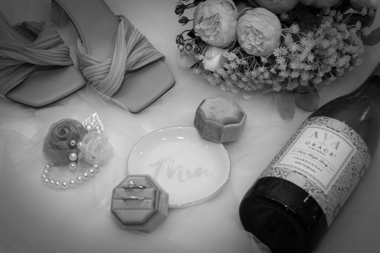 Black and white flat lay of wedding rings, jewelry box, wine bottle, and floral bouquet on white surface.