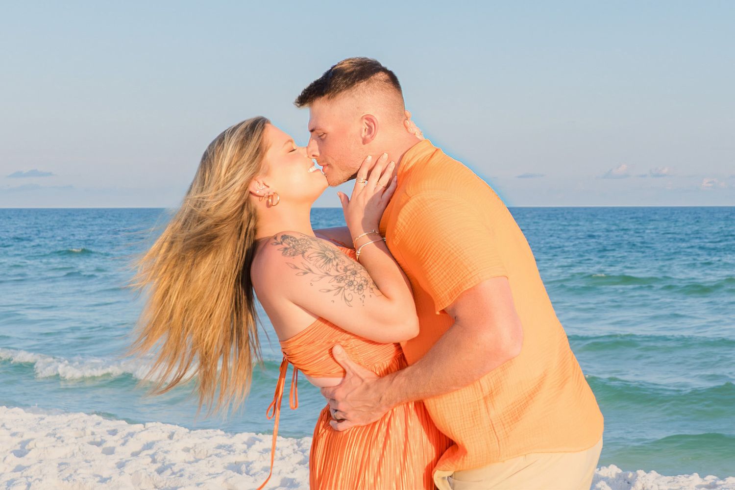 Couple sharing a romantic kiss on a sunny beach, wearing matching orange outfits with ocean waves in the background.