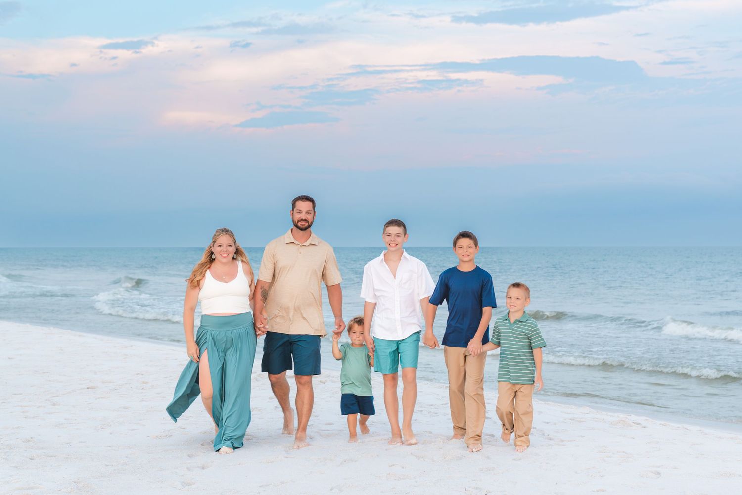 A family of six stands together on a beach at sunset, smiling and holding hands near the ocean waves.