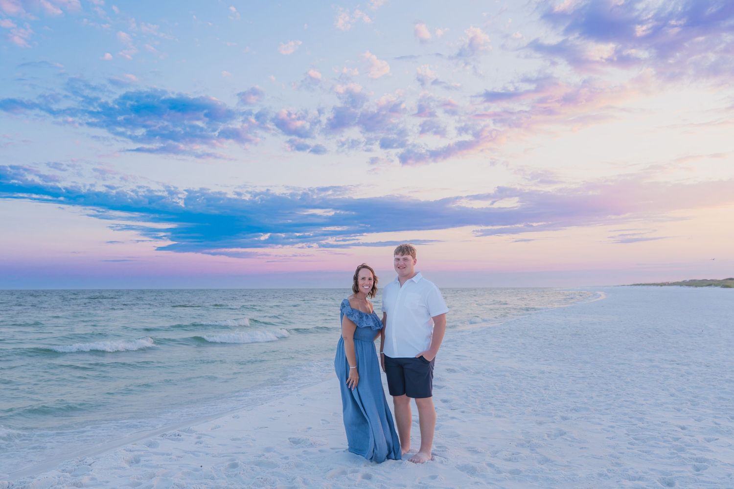 Couple posing on white sand beach at sunset with pastel pink and blue sky over calm ocean waters.