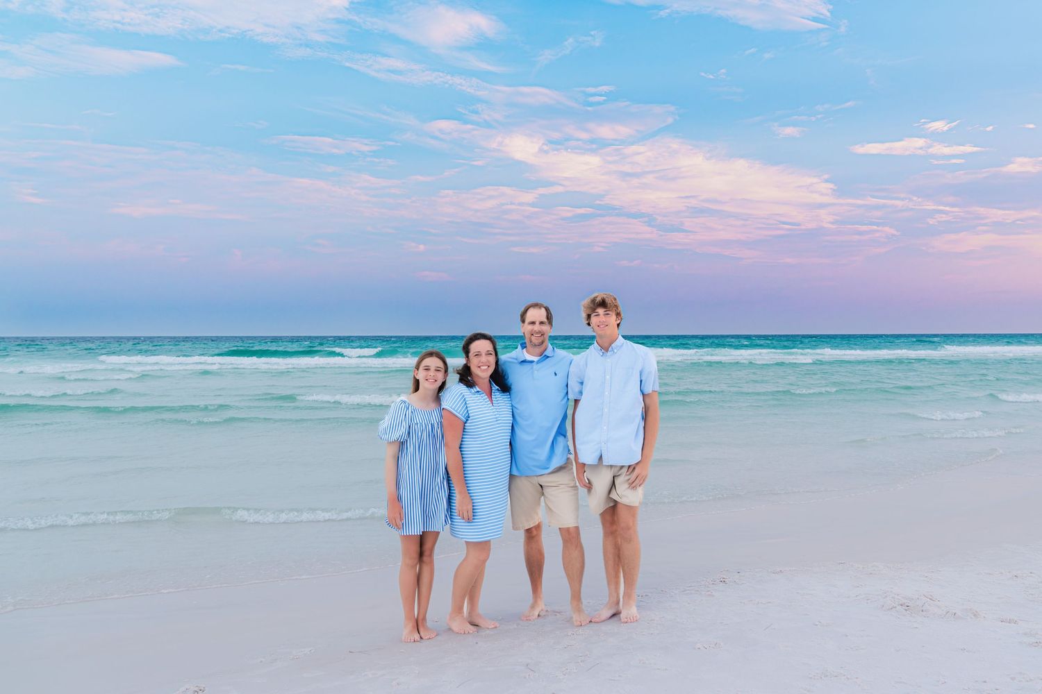Family of four in blue outfits posing on a white sand beach at sunset with turquoise water behind them.