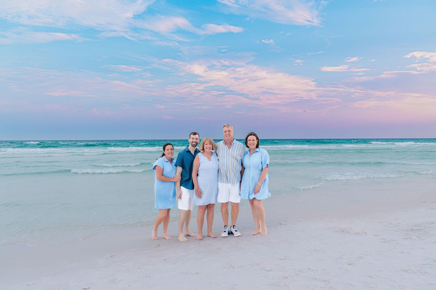Family of five posing on a white sand beach at sunset with pastel pink and blue sky over calm turquoise ocean.