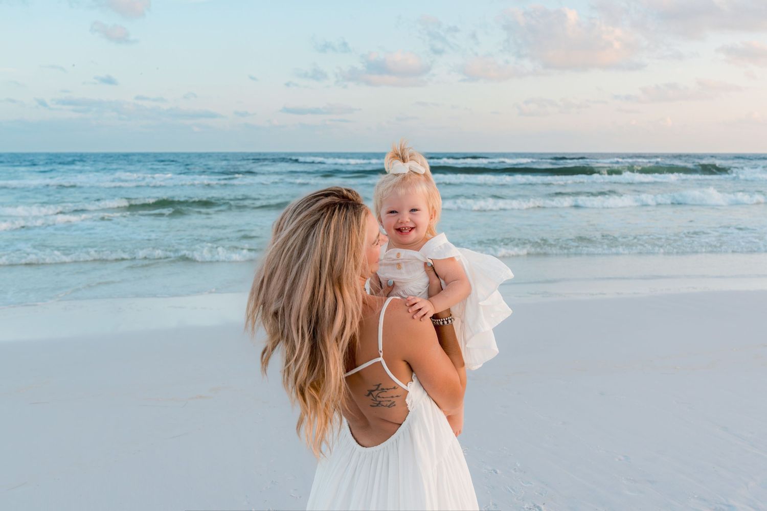 A mother in a white dress holds her smiling baby girl on a sandy beach with turquoise waves in the background.