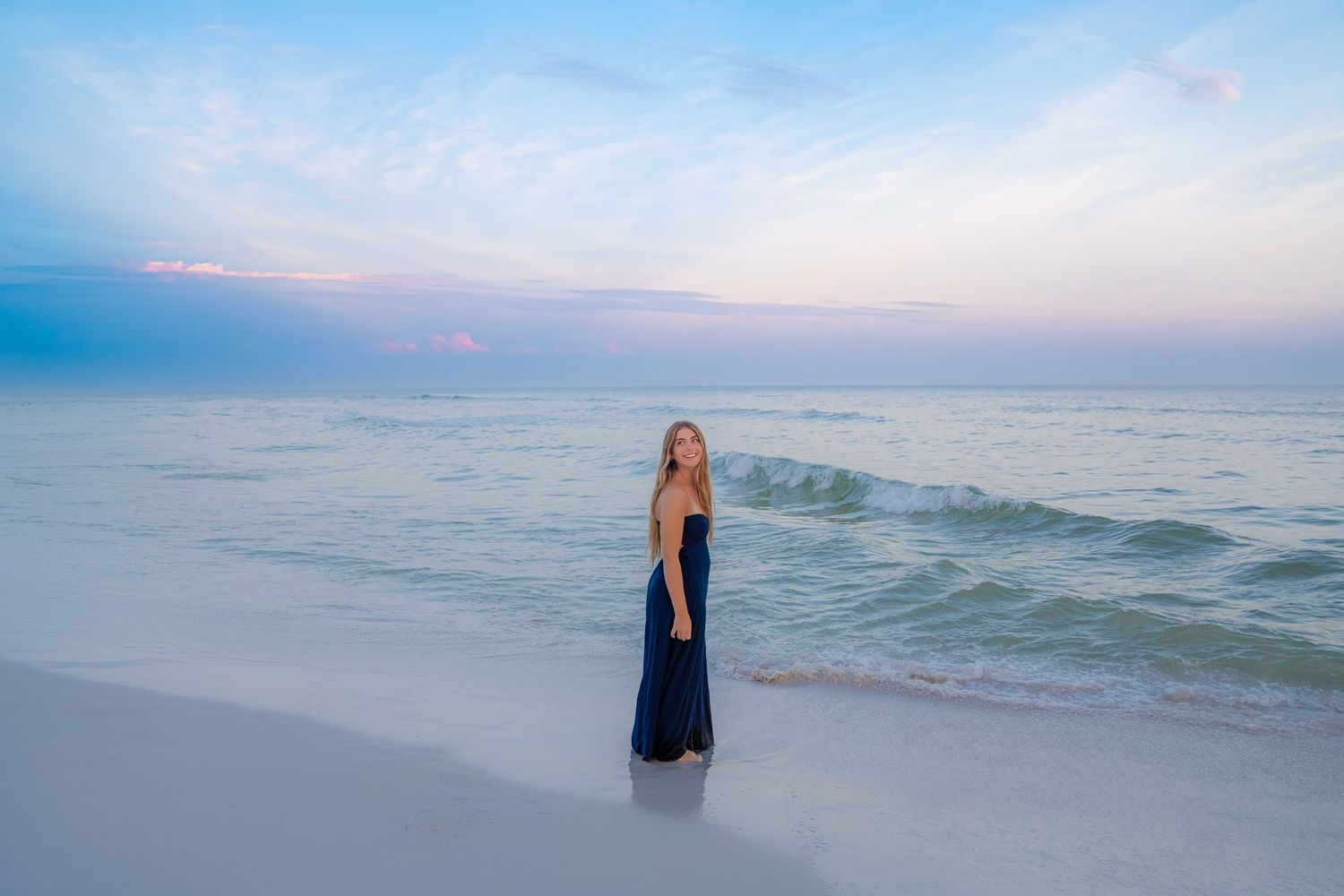 A woman in a navy maxi dress stands on a white sandy beach at sunset with gentle waves behind her.