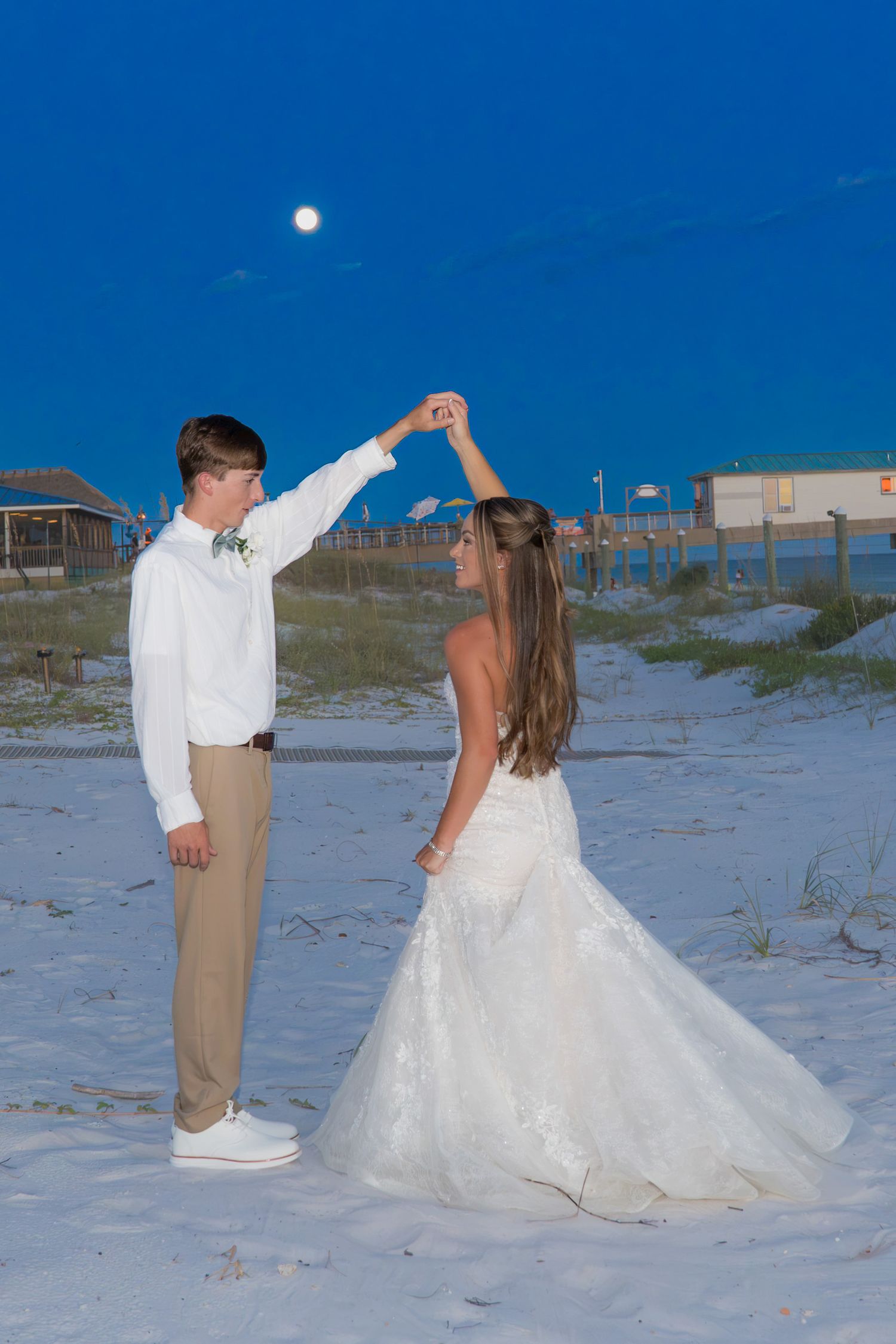 Bride and groom dancing on white sand beach at twilight with full moon, woman in white gown being twirled by man in white shirt.