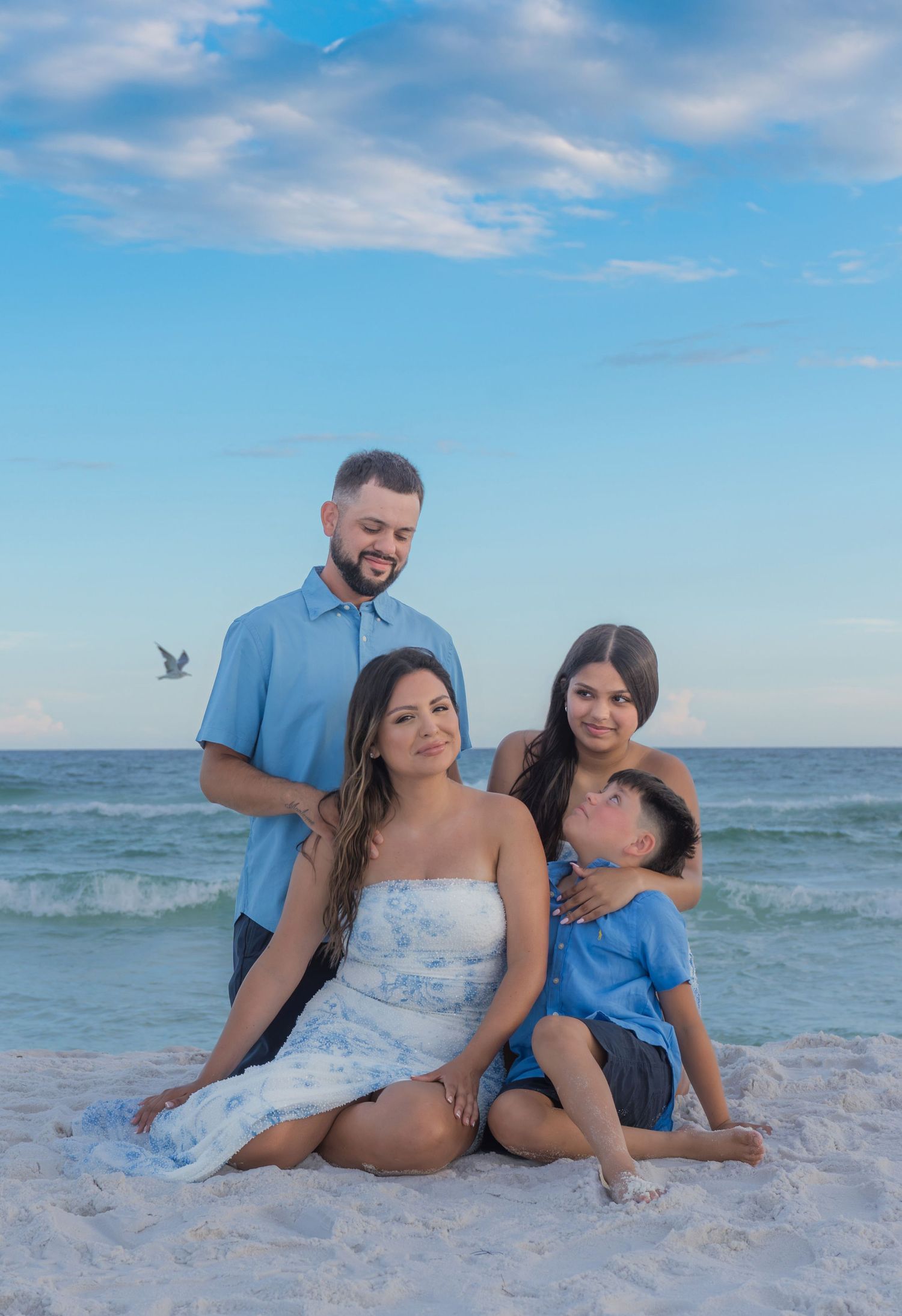 Family of four posing together on a sandy beach with turquoise ocean waves and blue sky backdrop.