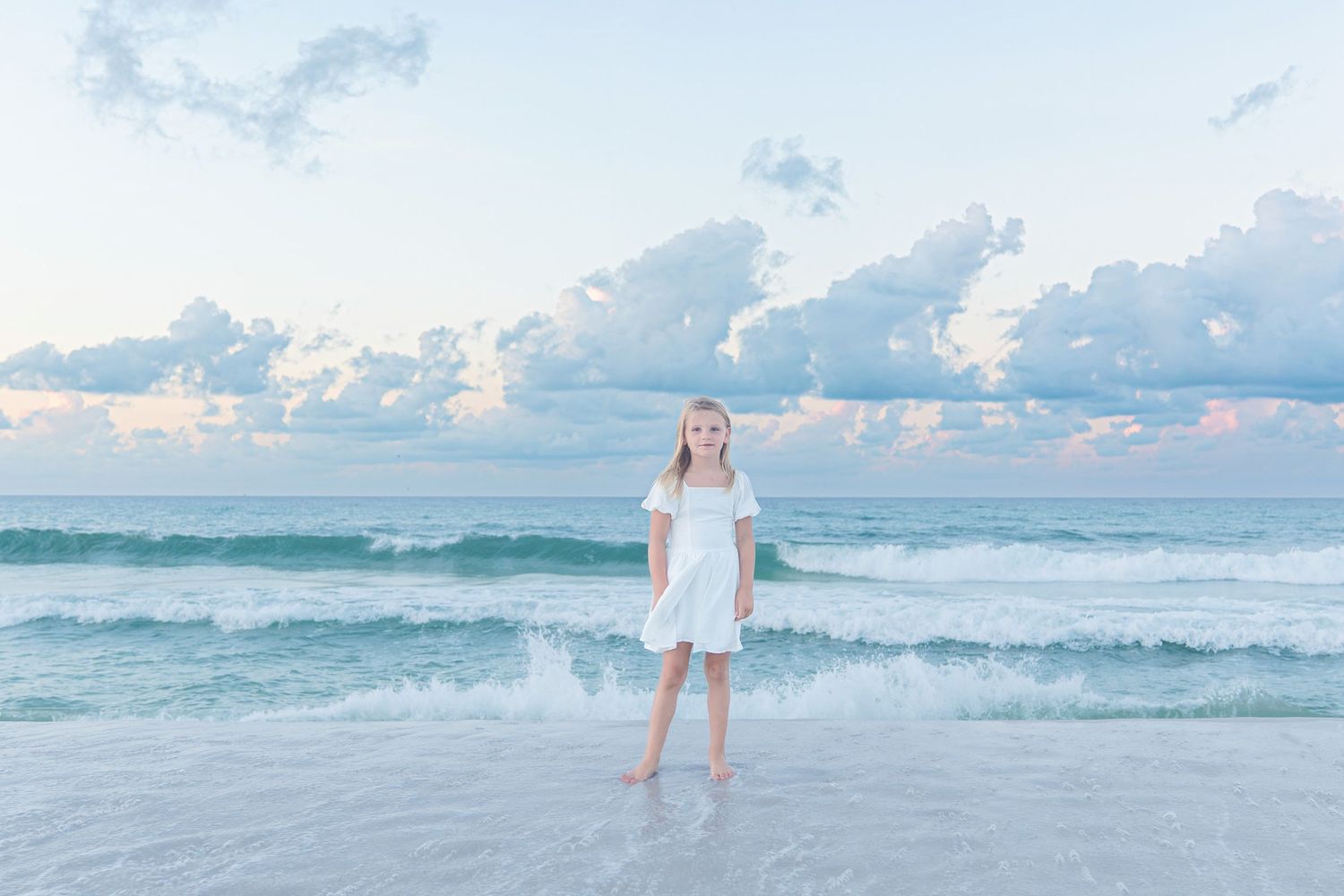 A young girl in a white dress stands barefoot on a sandy beach with turquoise waves and pastel cloudy sky.