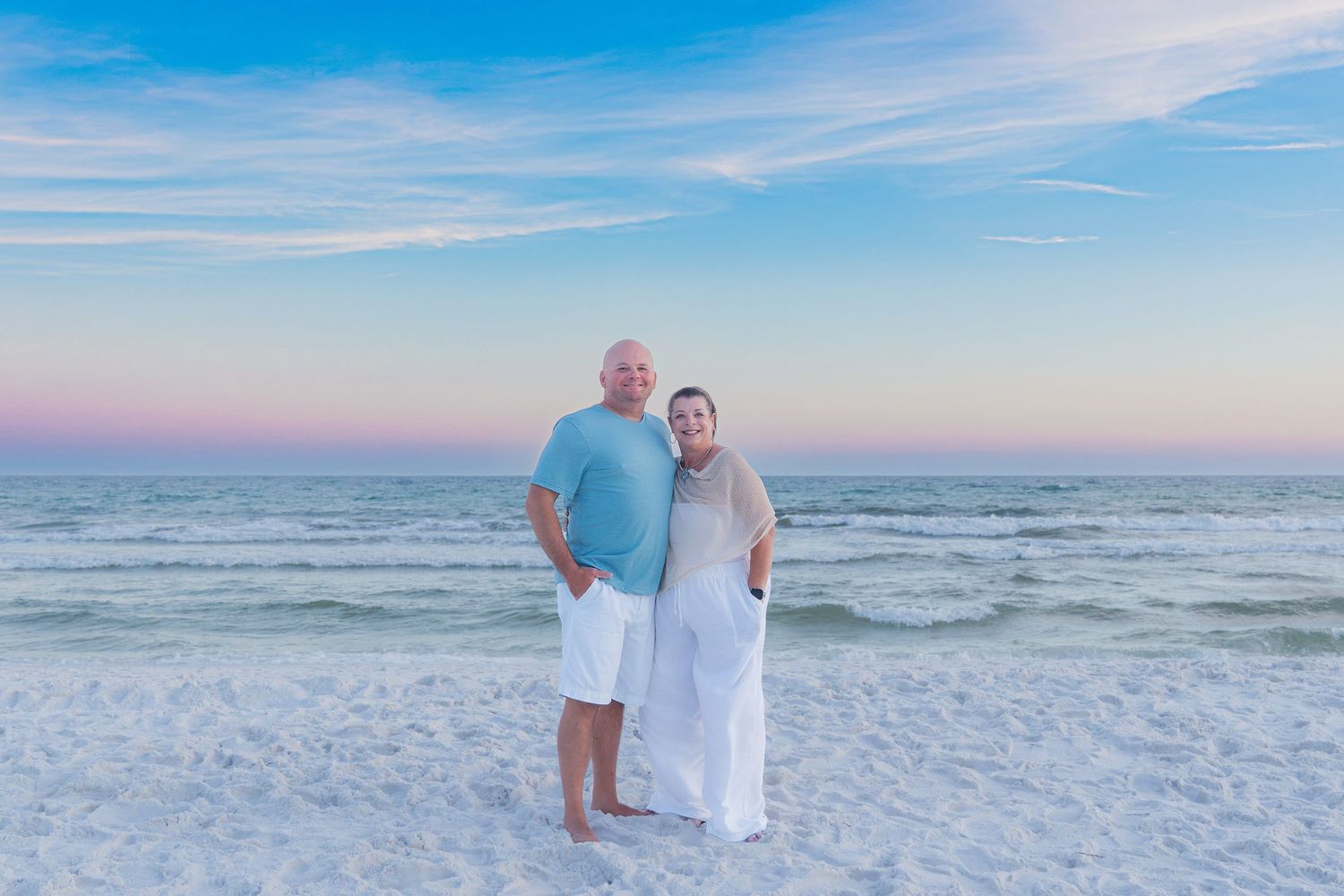 Couple smiling together on a white sand beach at sunset with calm turquoise ocean waves in the background.
