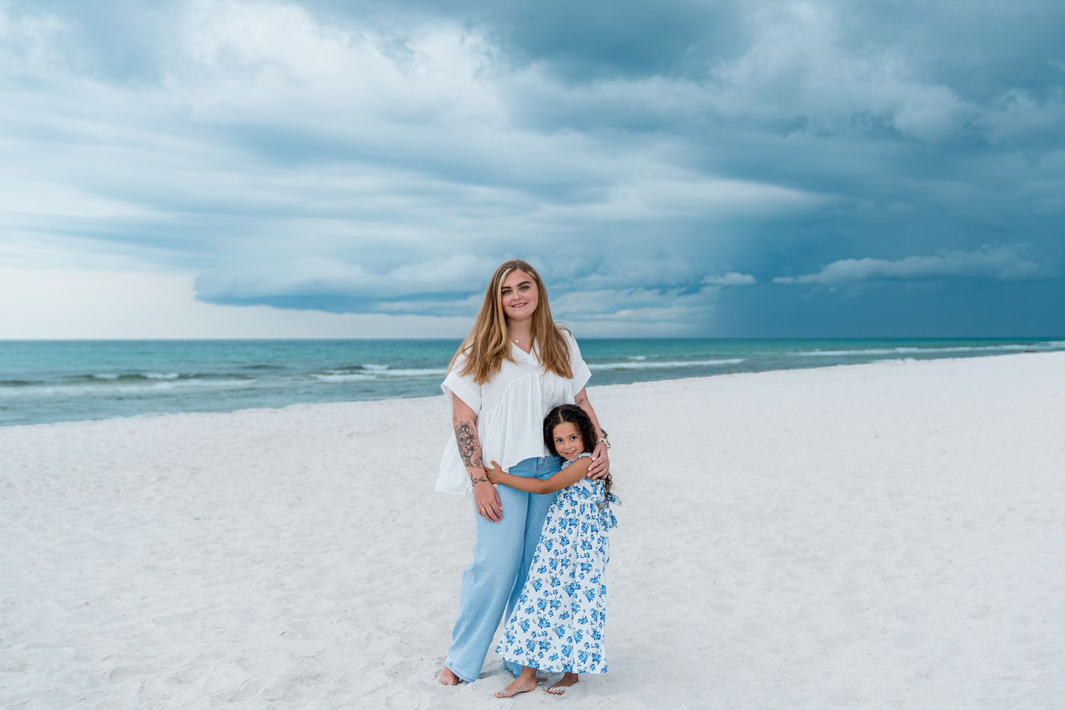 A mother and daughter pose on a white sandy beach under dramatic storm clouds near the ocean.