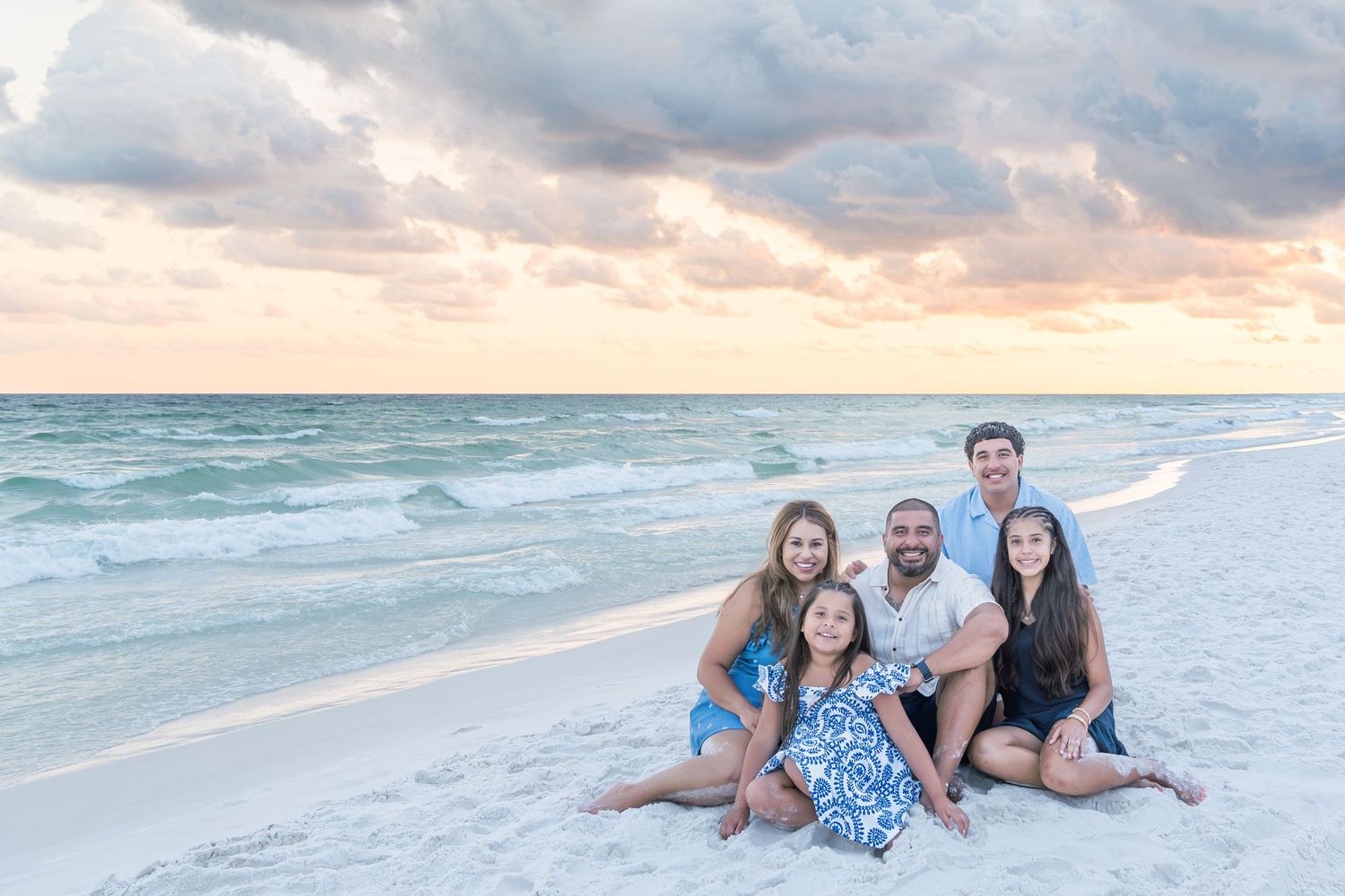 Family of five smiling together on a white sandy beach at sunset with colorful clouds and turquoise ocean waves.