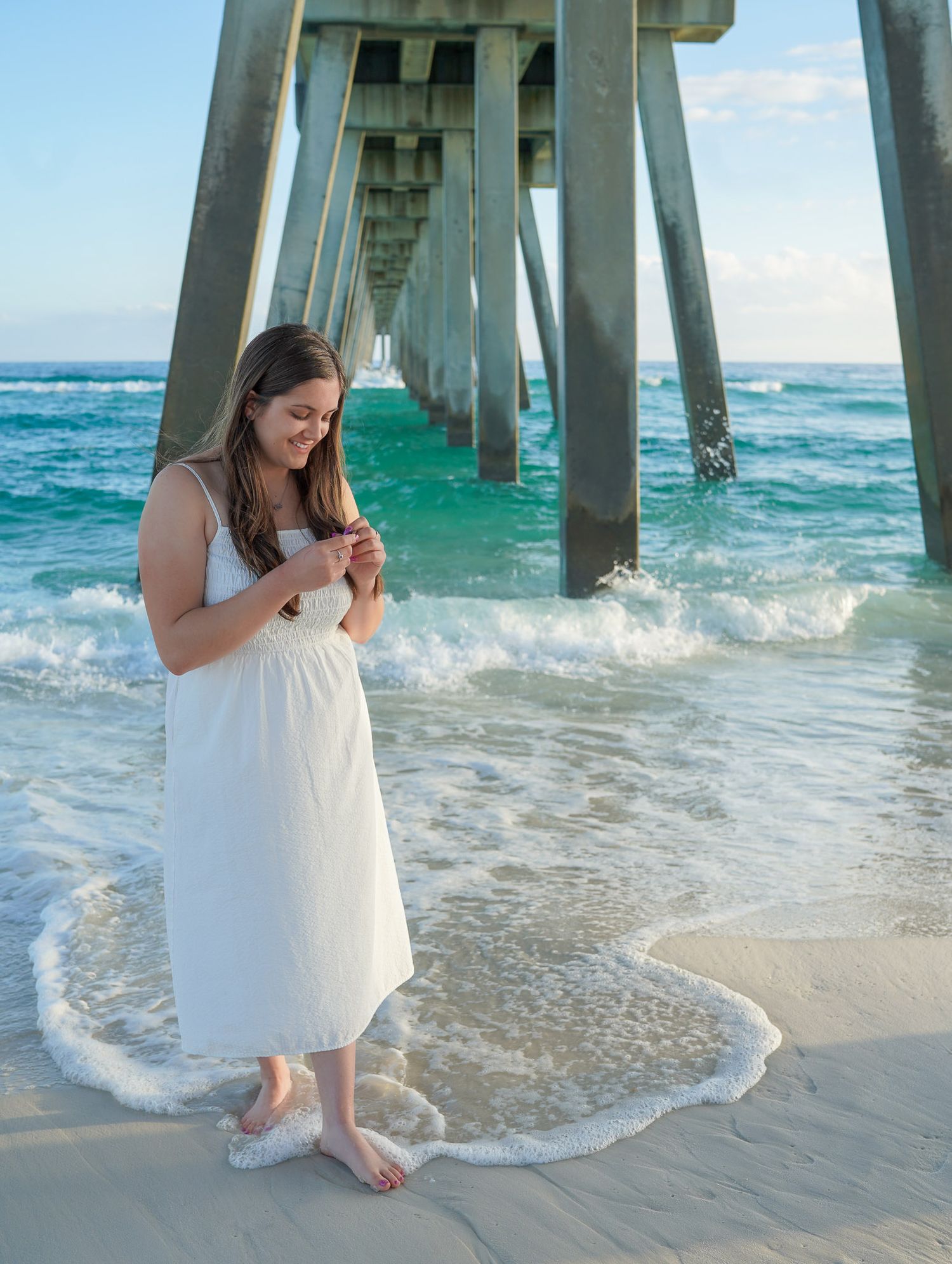 Woman in white sundress stands barefoot on sandy beach beneath a large ocean pier at sunset.