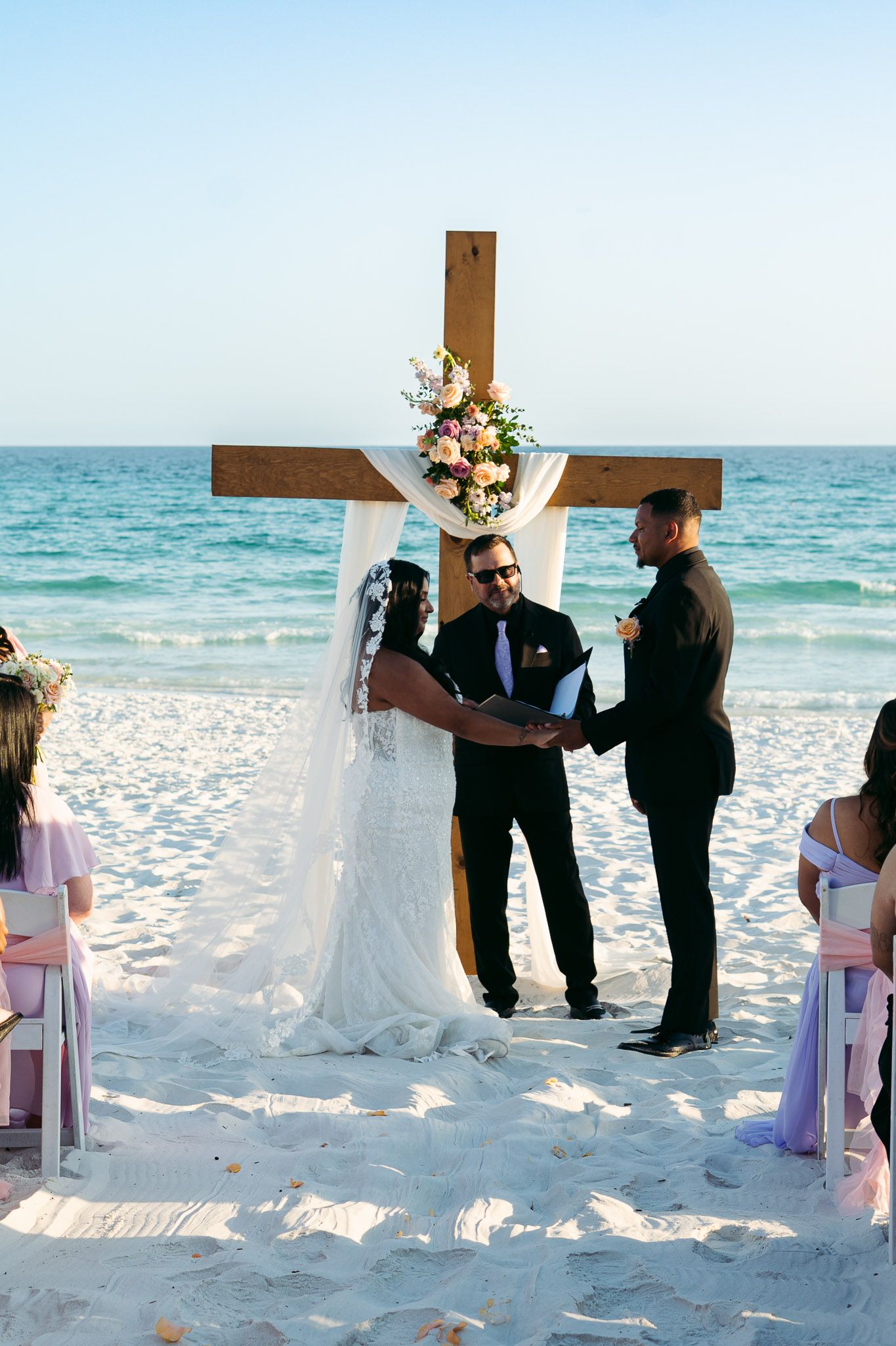 Bride and groom exchanging vows at a beach wedding ceremony before a wooden cross altar.