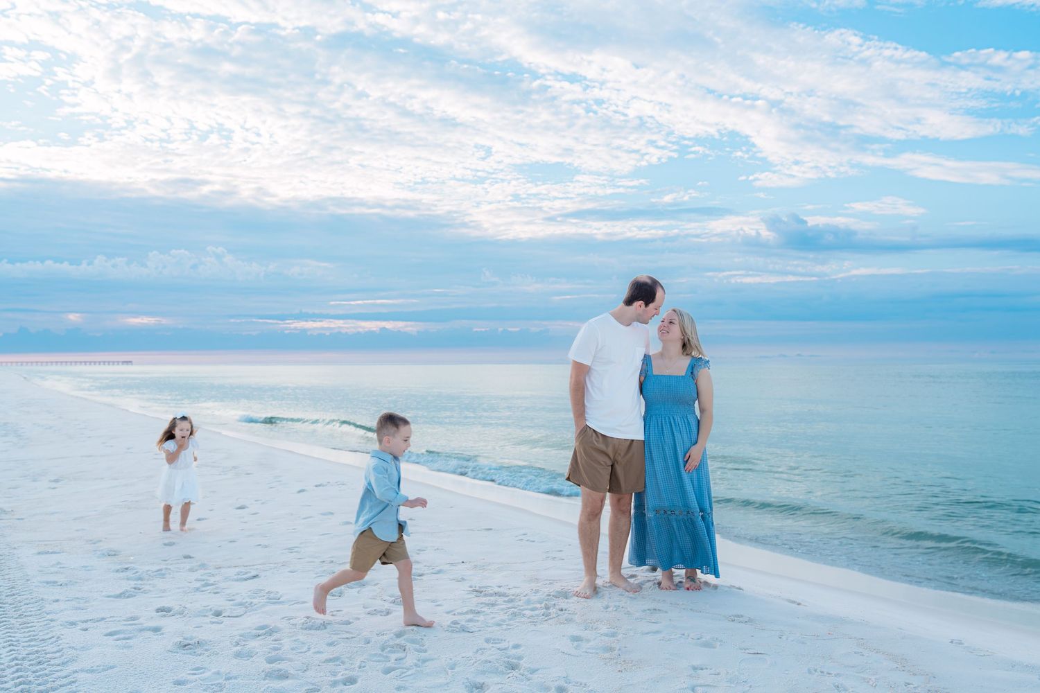 Family of four enjoying a serene beach at dusk, with two children playing near the shoreline and calm blue waters.
