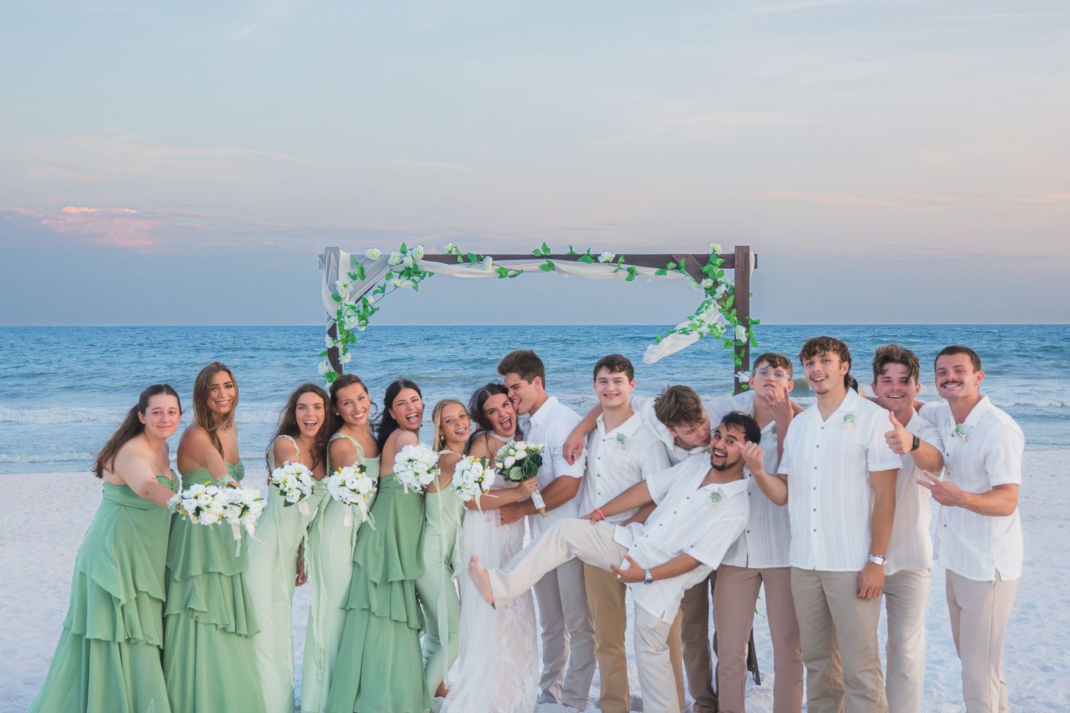 Wedding party poses on beach at sunset with green bridesmaid dresses and floral arch backdrop.