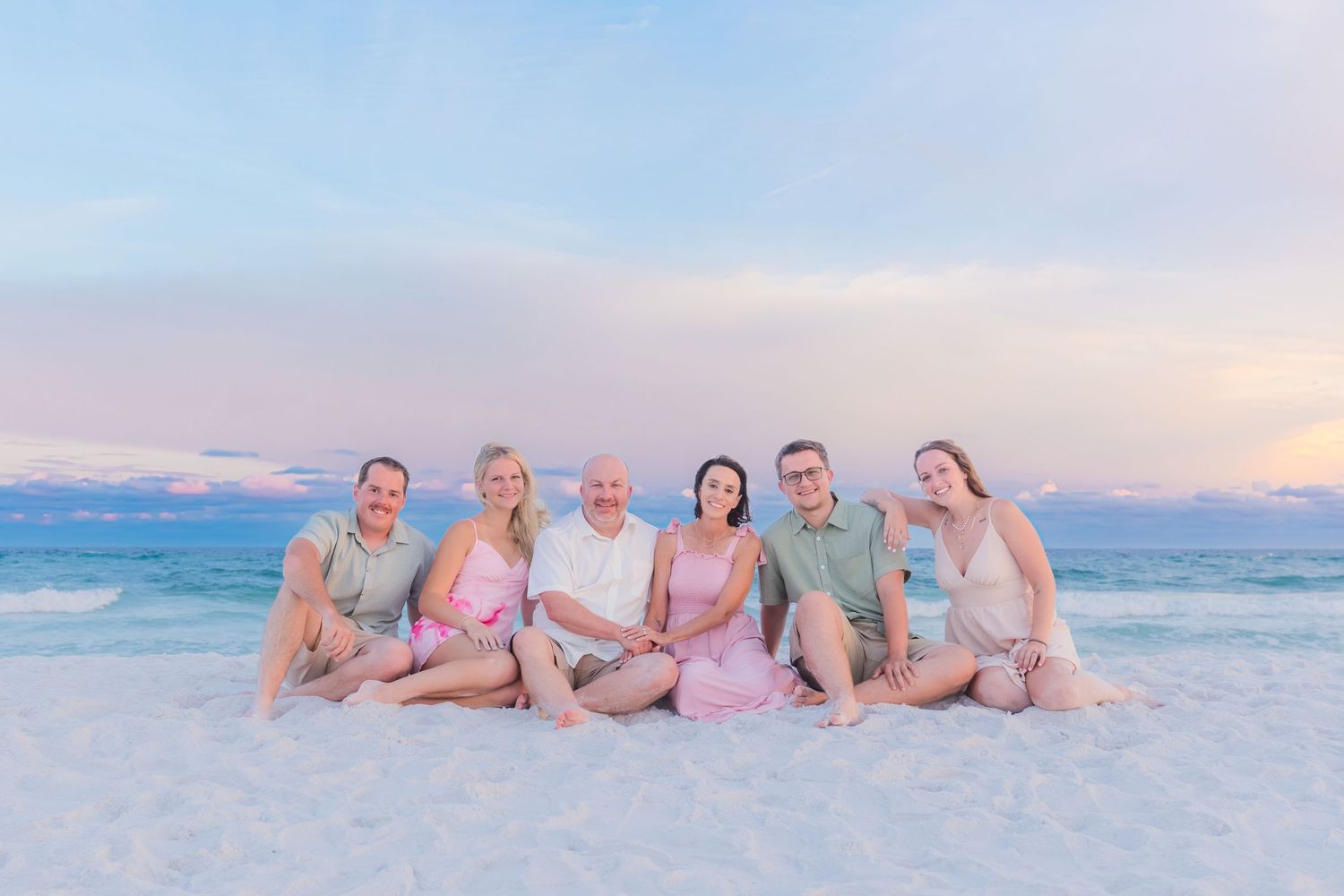 A group of seven adults sit together smiling on a white sand beach at sunset.