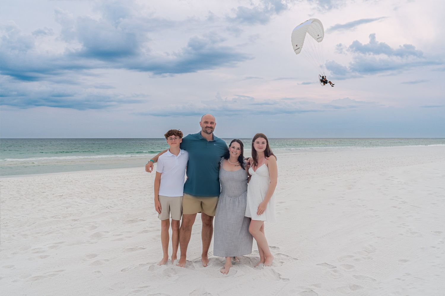 Family of four smiling together on a white sand beach with calm ocean waves and a paramotor flying in the background.