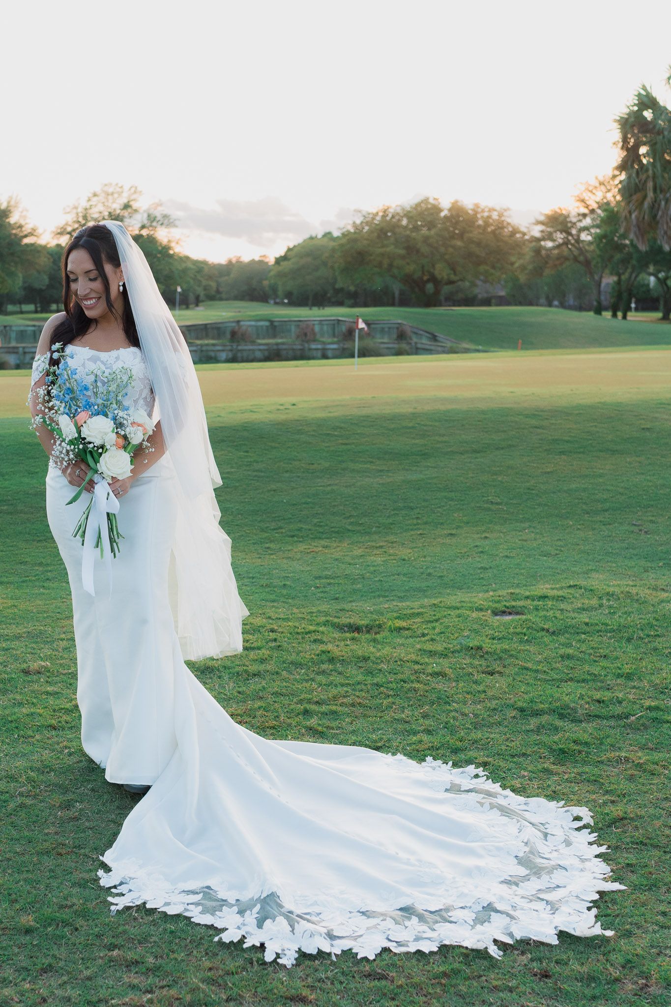 Bride in white gown with floral train and veil holds bouquet on green golf course at sunset.