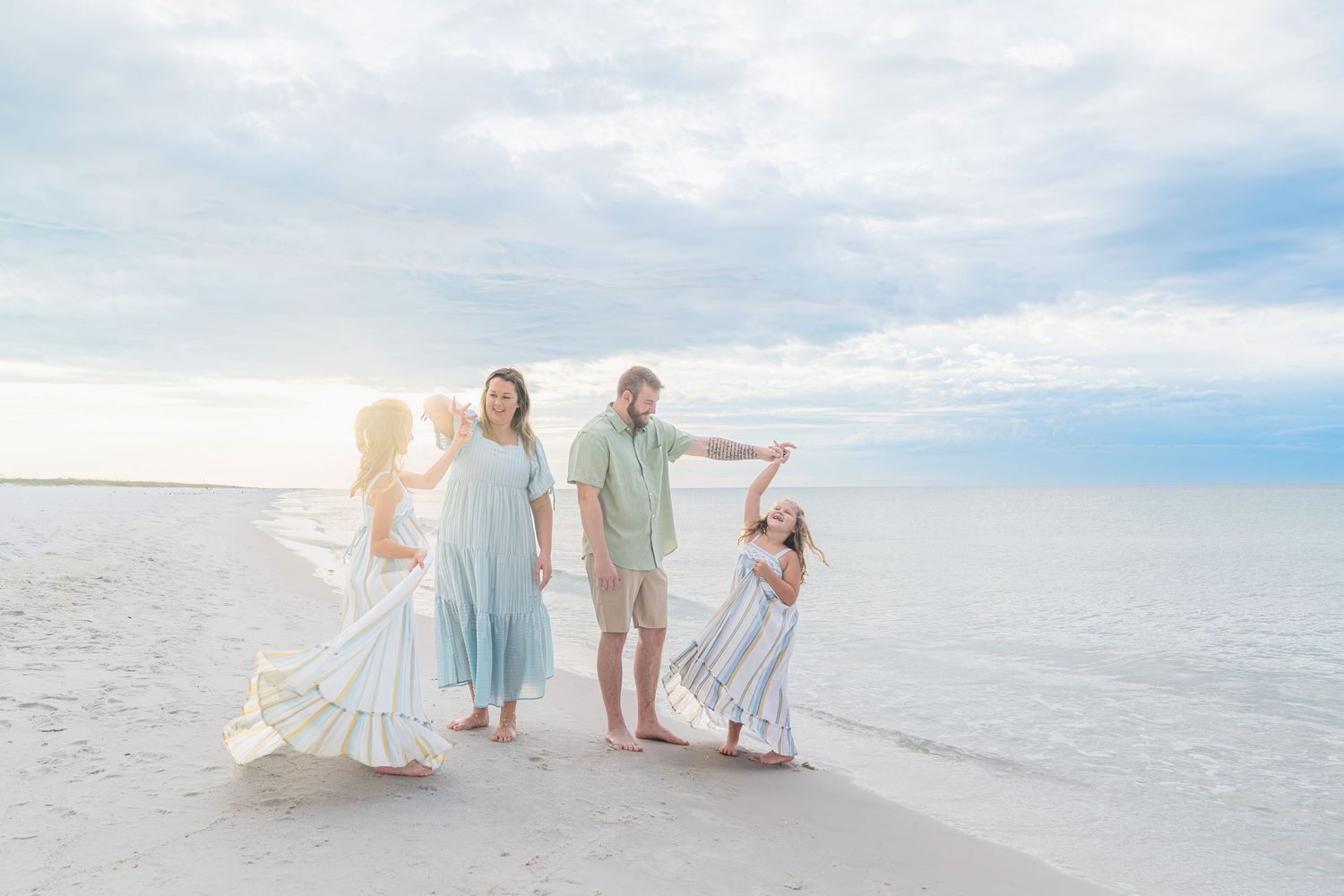 Happy family of four posing together on a white sand beach under a cloudy blue sky.