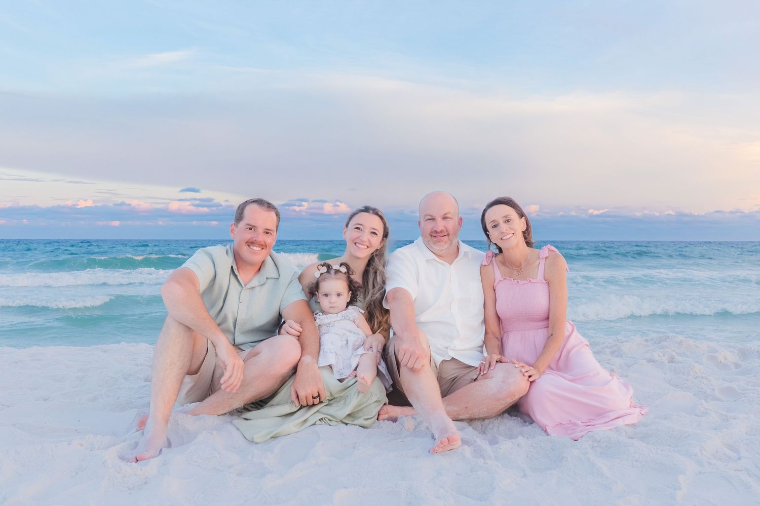 Family of five sits together on white sand beach smiling with turquoise ocean waves and pastel sunset behind them.