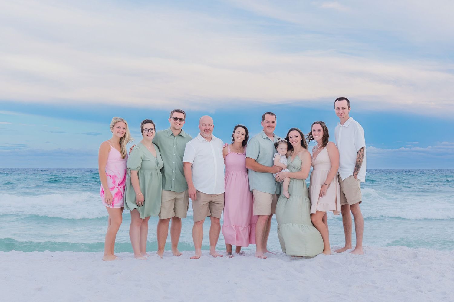 A large family group poses together on a sandy beach at sunset, dressed in coordinated pastel outfits.