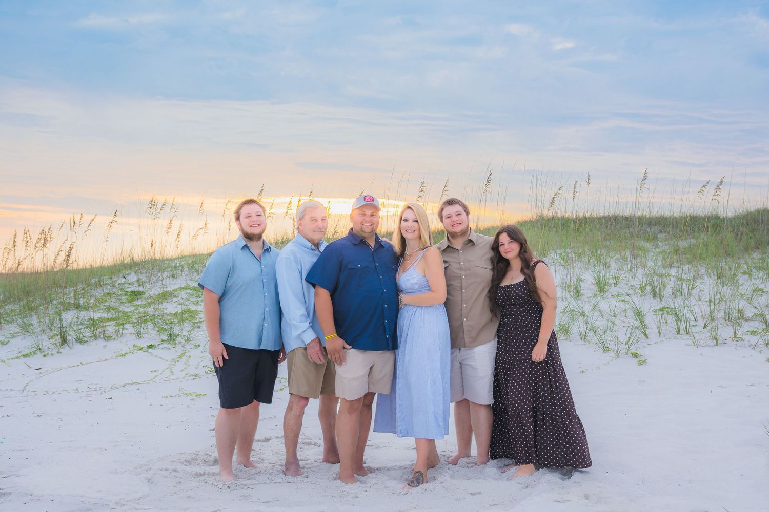 Family of six posing together on a sandy beach at sunset with sea oats and pastel sky in the background.