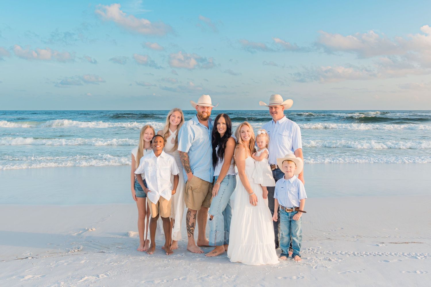 Family of seven posing together on a white sand beach with turquoise ocean waves in the background.
