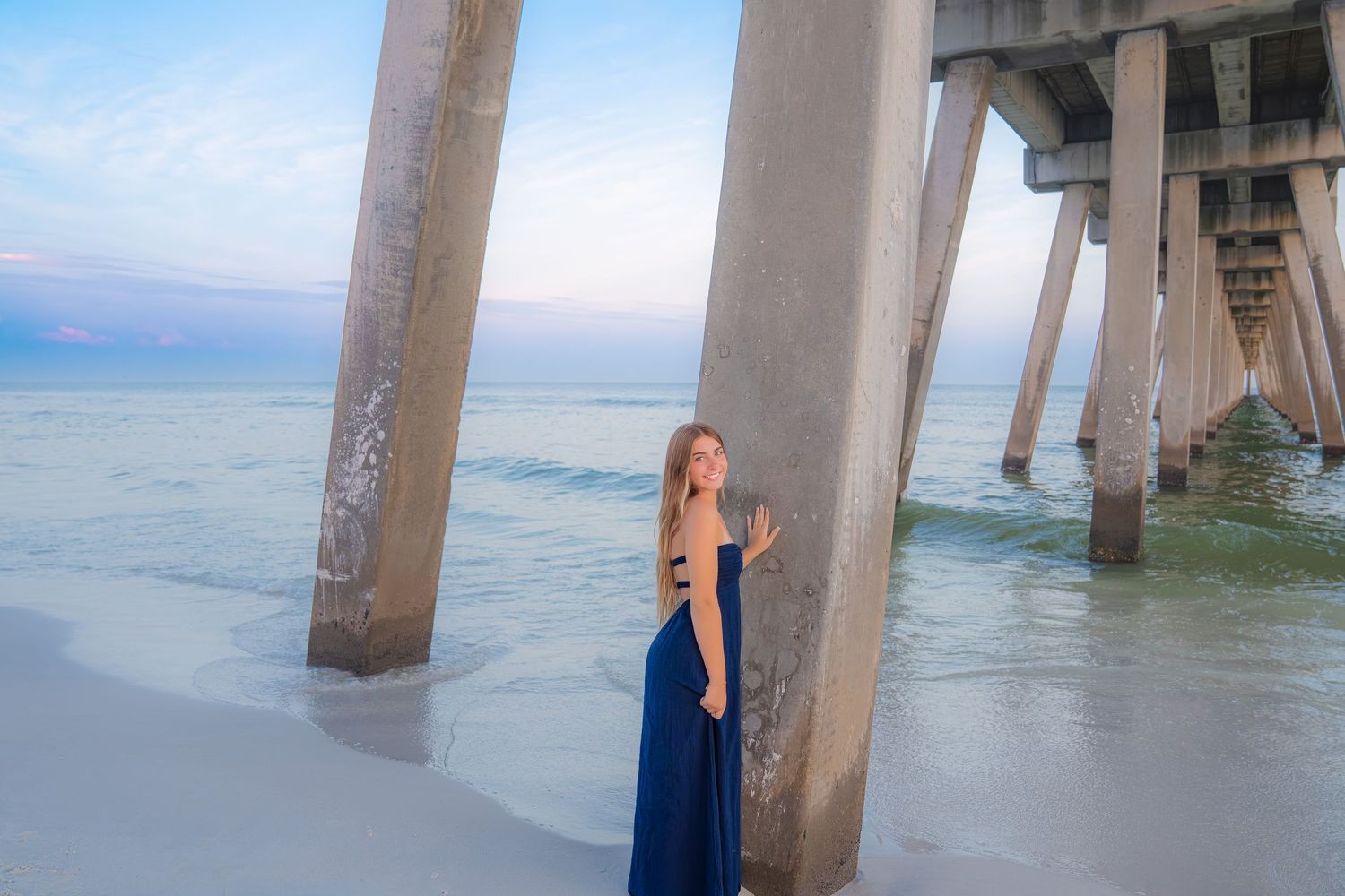 Woman in blue maxi dress poses under a pier on a white sand beach at dusk with calm ocean waves.
