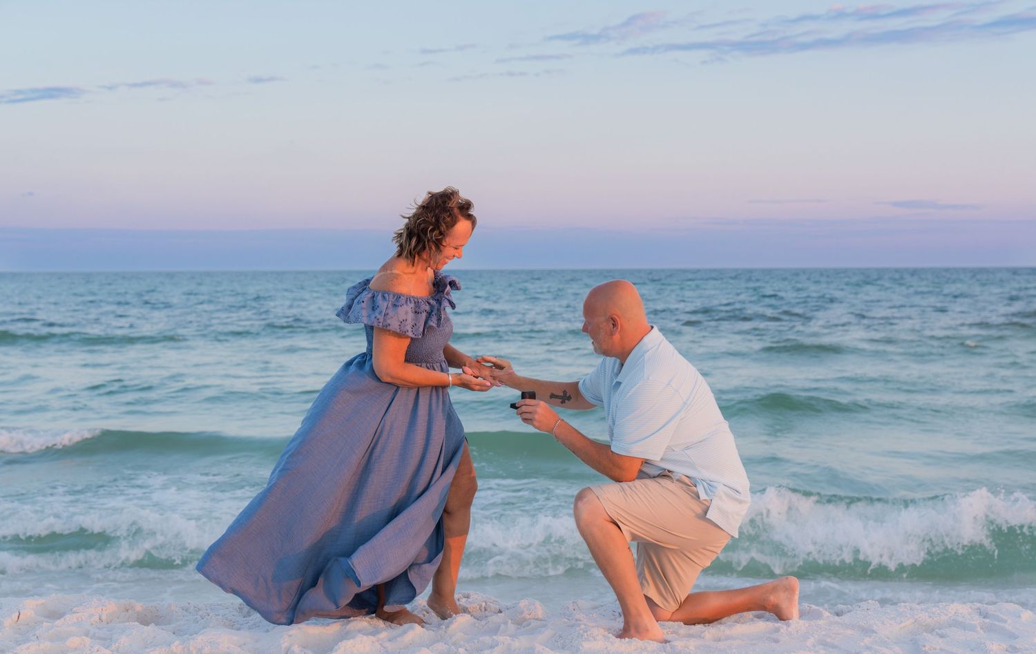 Man proposes to woman on a sandy beach at sunset, waves crashing behind them, woman in flowing blue gown.