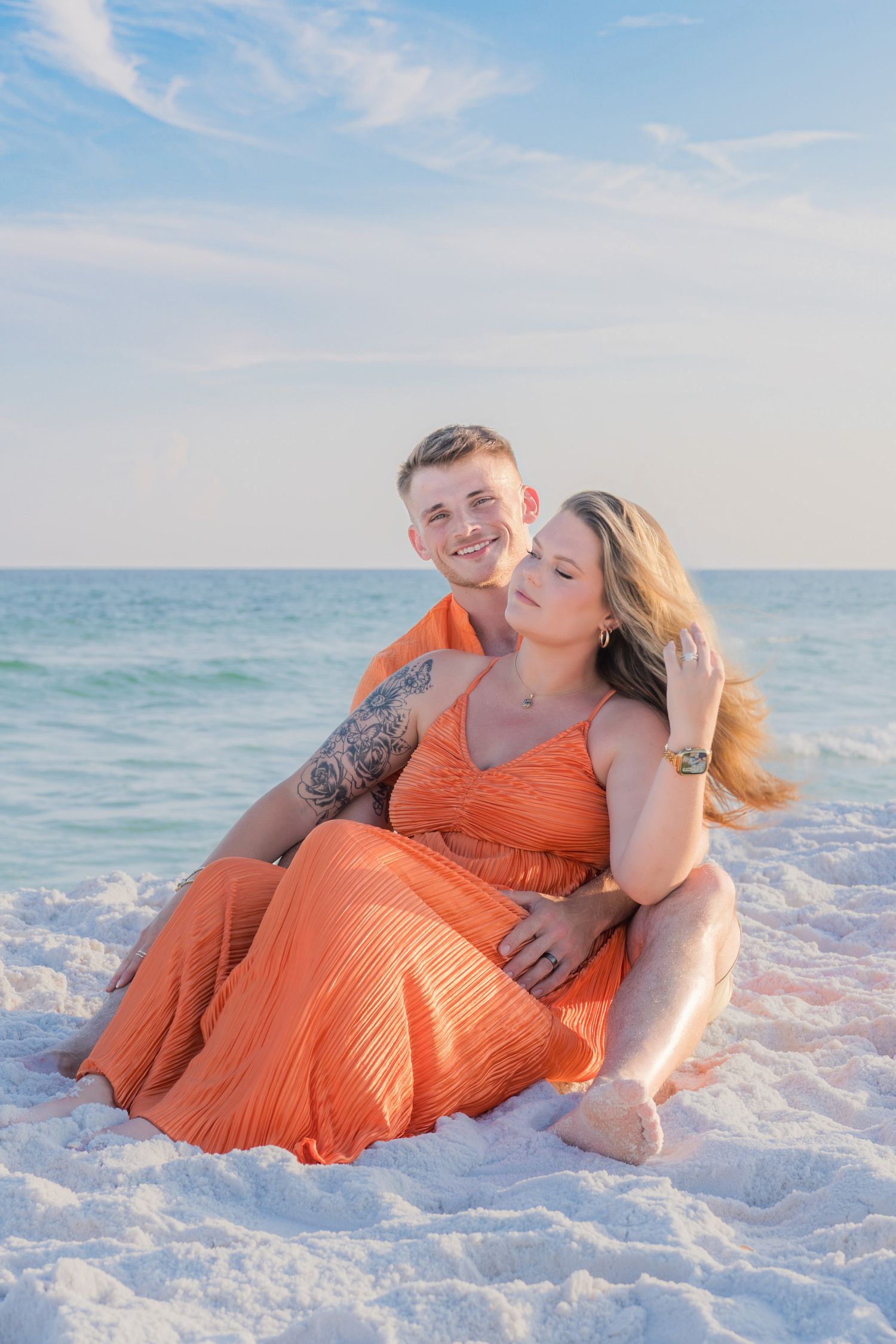 Couple in matching orange outfits sitting together on white sand beach with turquoise ocean backdrop.