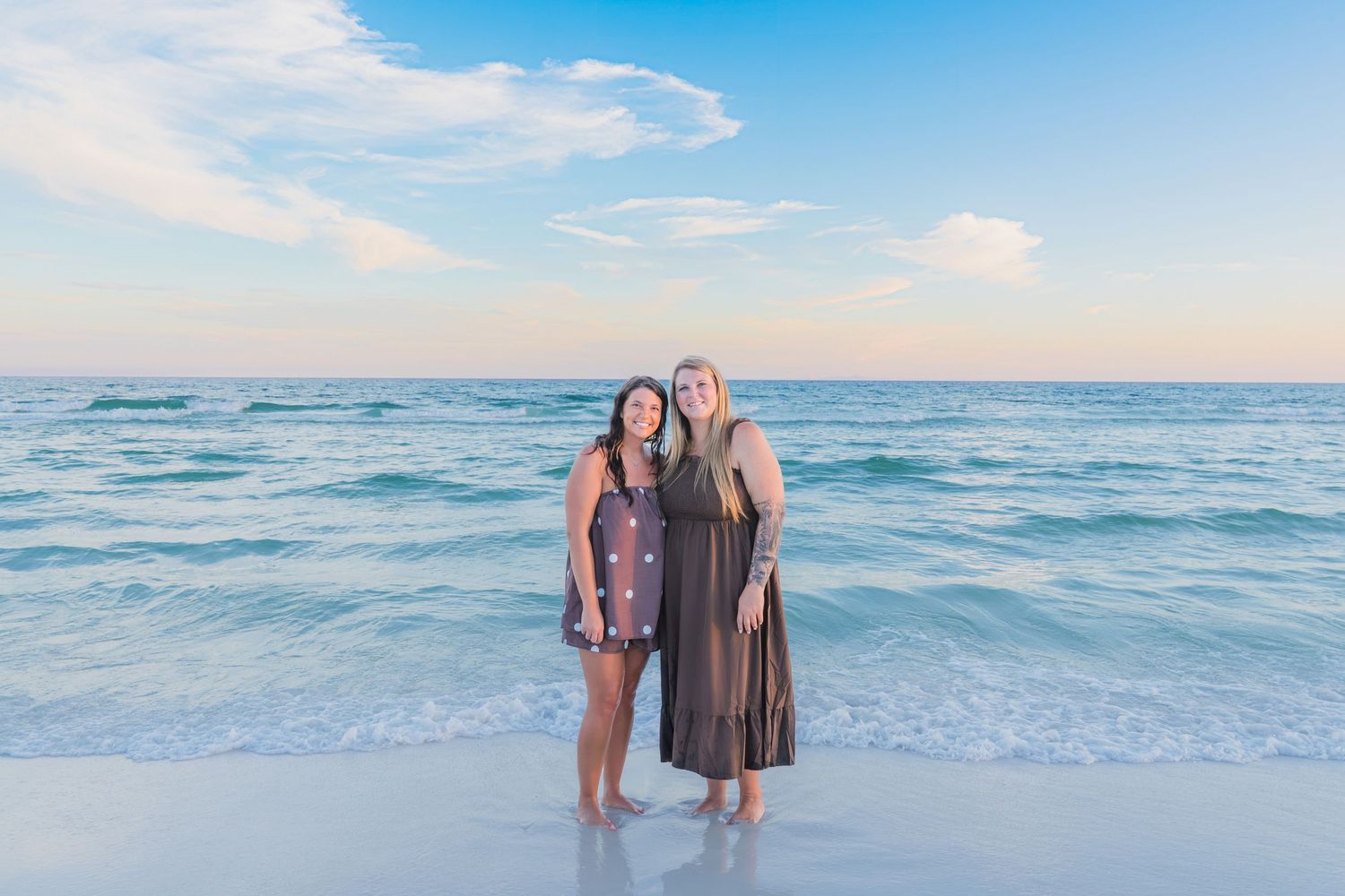 Two women smiling together on a sandy beach with turquoise ocean waves and blue sky behind them.