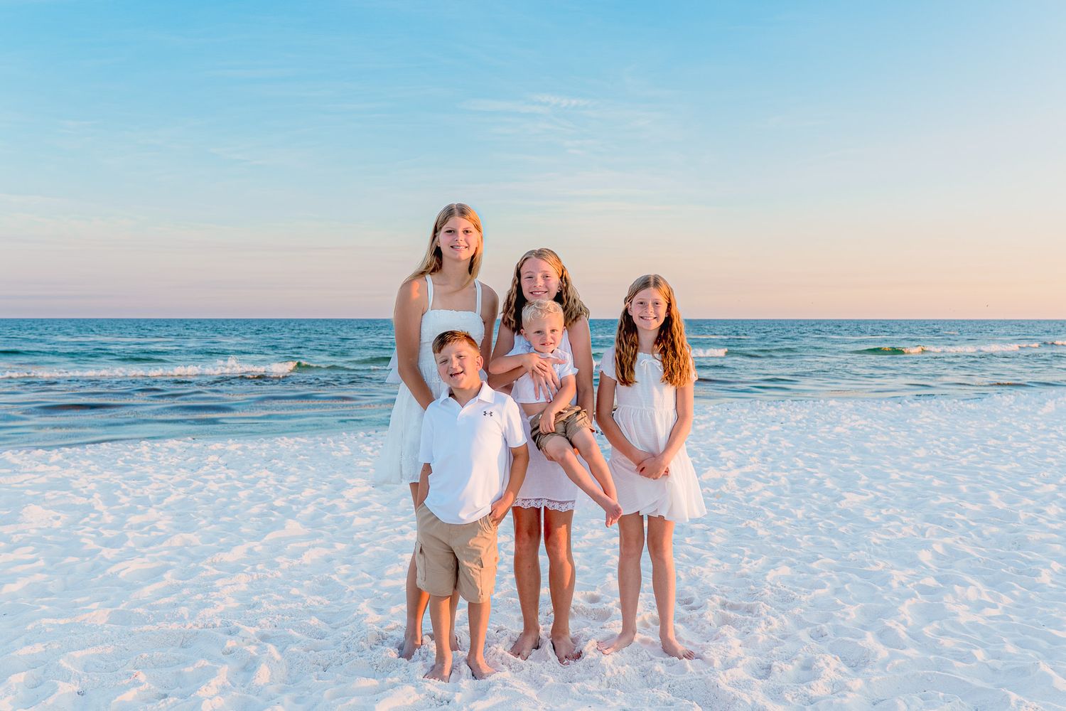 Four children pose together on a white sand beach at sunset with calm ocean waves in the background.