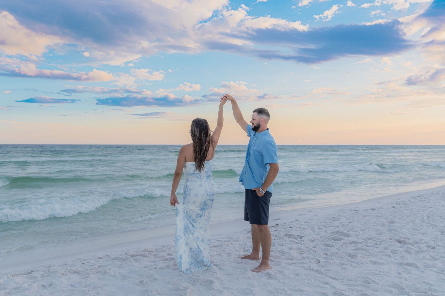 Couple dances on white sand beach at sunset, man spinning woman in flowing white dress by turquoise water.