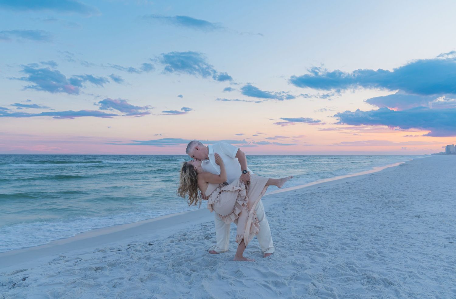 Couple sharing a romantic dip kiss on a white sand beach at sunset with pastel pink and blue skies.