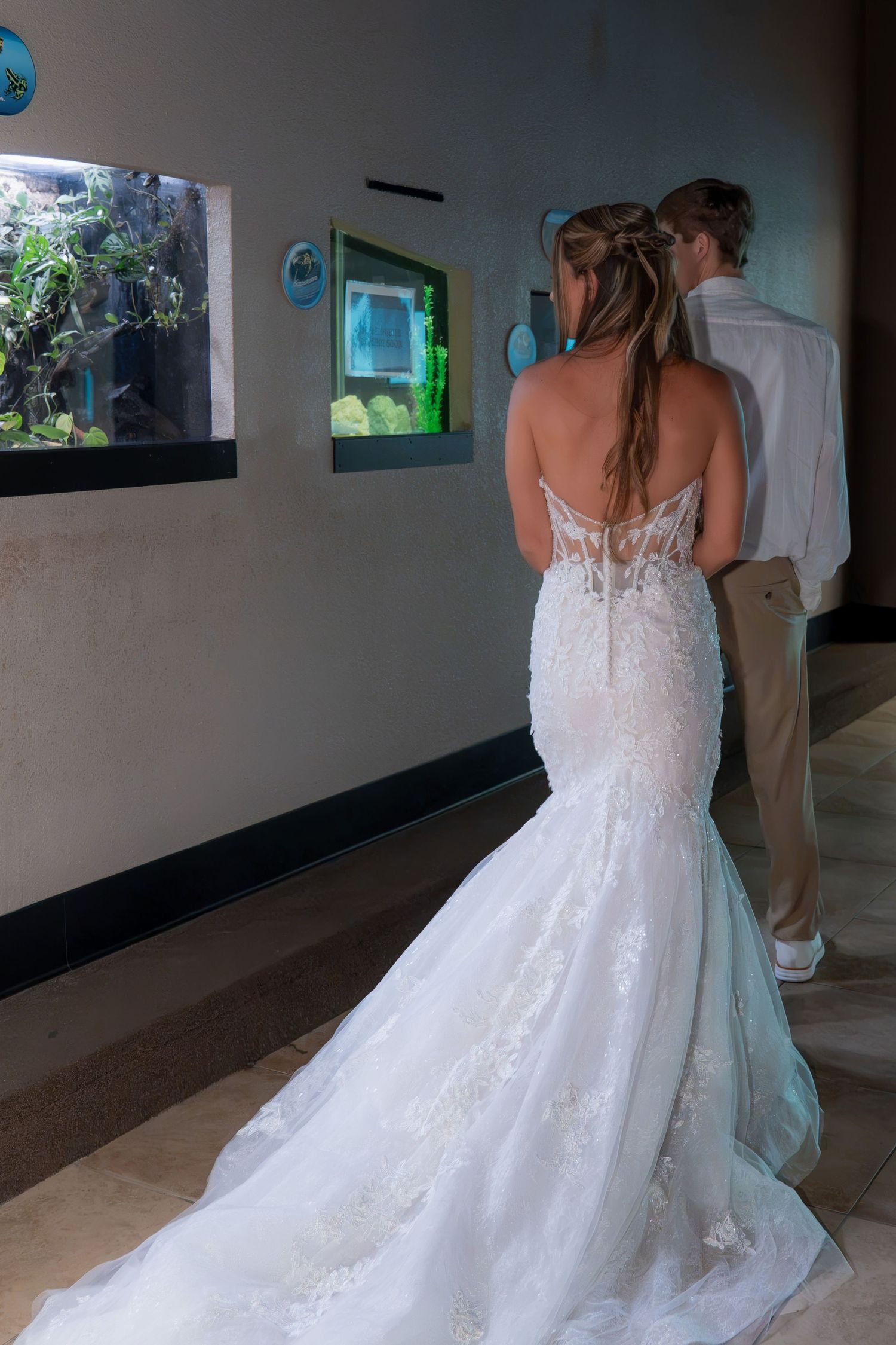 Bride in backless lace mermaid wedding dress viewing aquarium display with groom beside her.